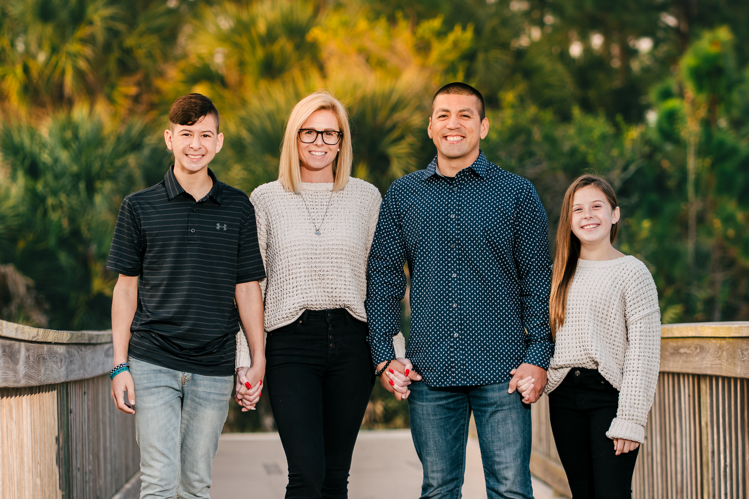 Family of four smiling together on a wooden bridge surrounded by lush green trees in warm natural light.