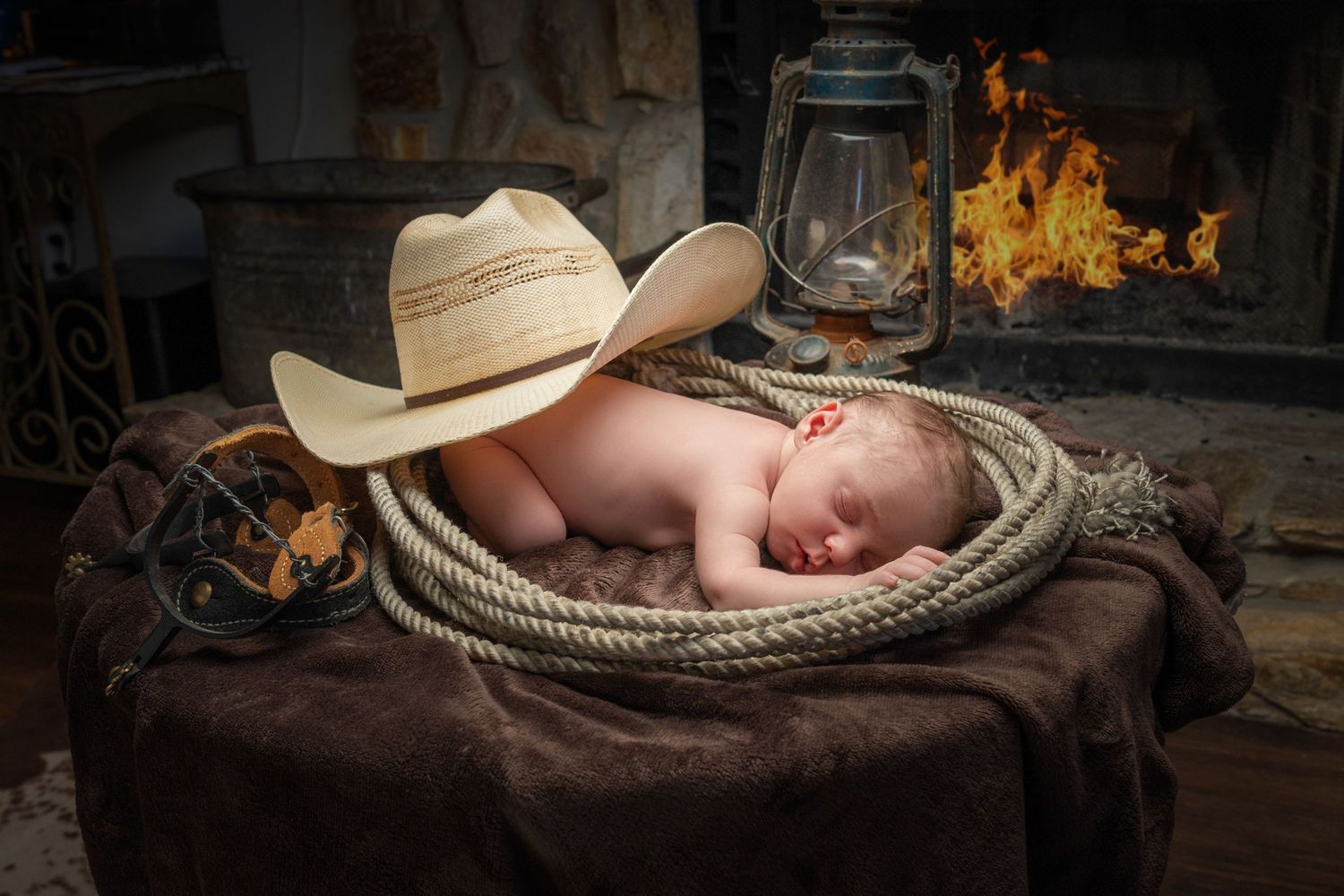 Newborn baby sleeping in a cowboy hat surrounded by rope near a fireplace with a lantern in a rustic western setting.