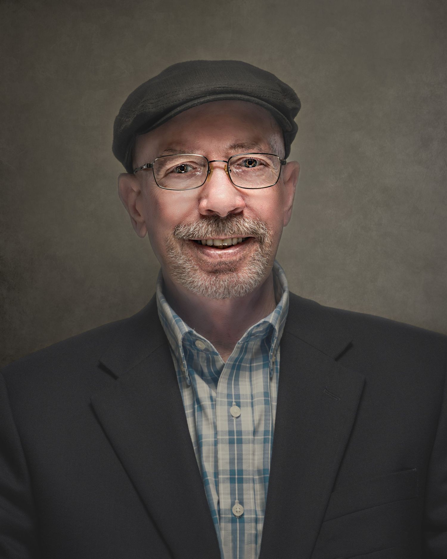 Smiling middle-aged man wearing glasses, flat cap, plaid shirt, and dark blazer poses for a professional portrait photo.