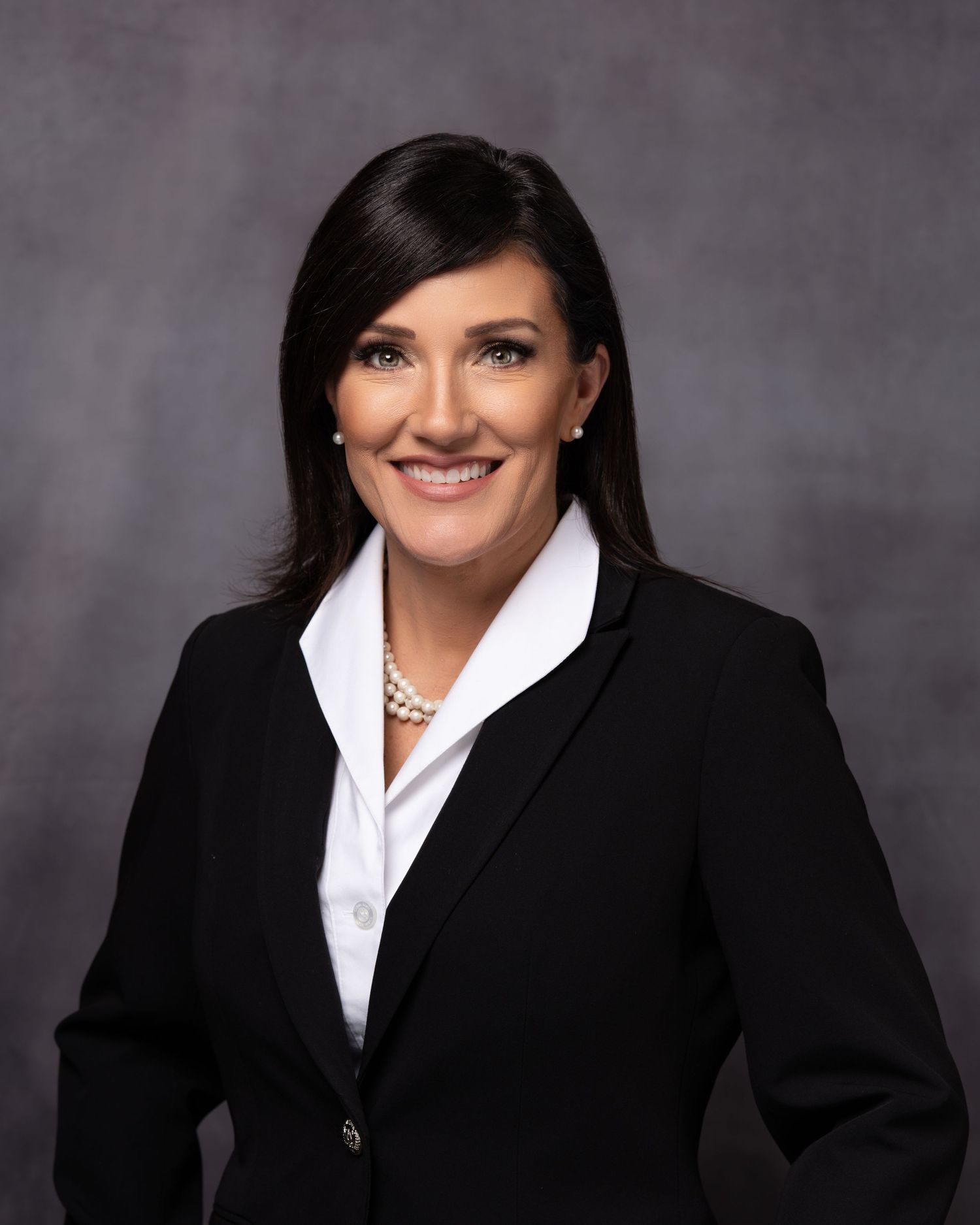 Professional headshot of a woman in a black blazer and pearl necklace smiling against a gray background.