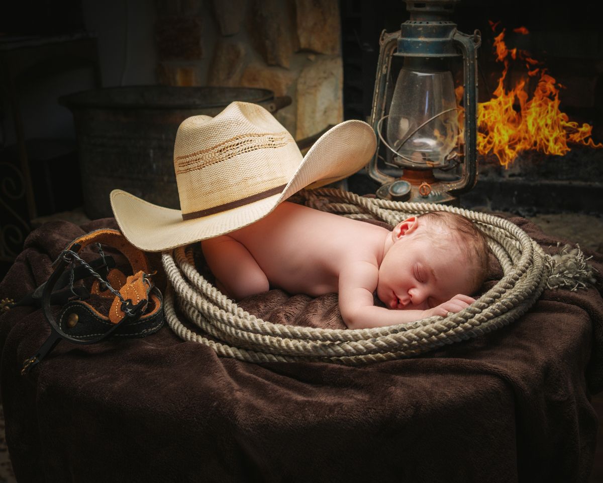 Newborn baby sleeping in a coiled rope with a cowboy hat beside a glowing lantern and fireplace.