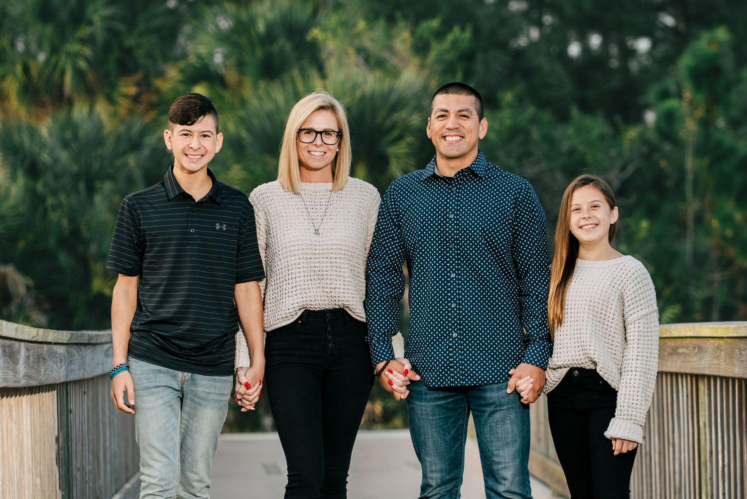 Family of four smiling together on a wooden bridge surrounded by lush green trees.