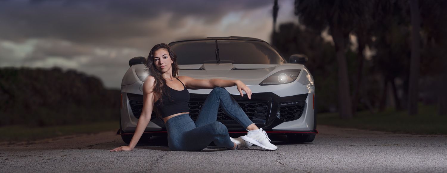 A woman in jeans and white sneakers poses in front of a sleek silver sports car on a dramatic cloudy evening.