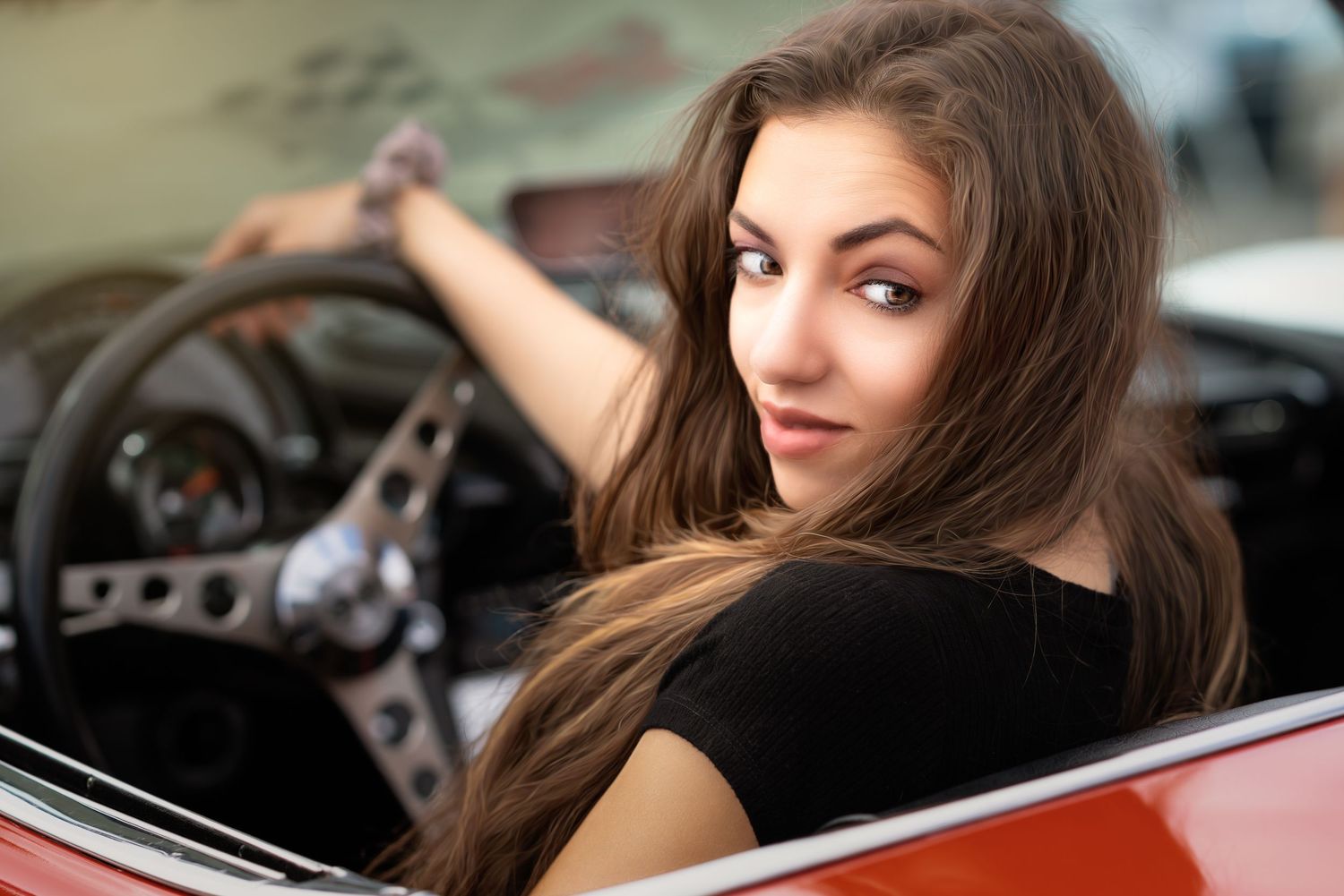 Young woman with long brown hair sitting in a classic red convertible car, looking over her shoulder.