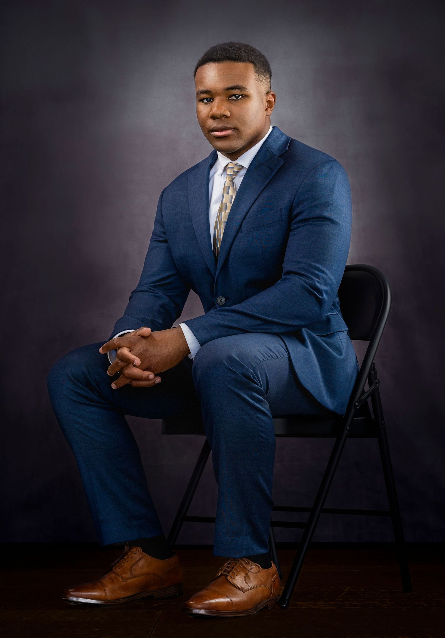 Young man in blue suit and tan dress shoes seated on folding chair against dark studio background.