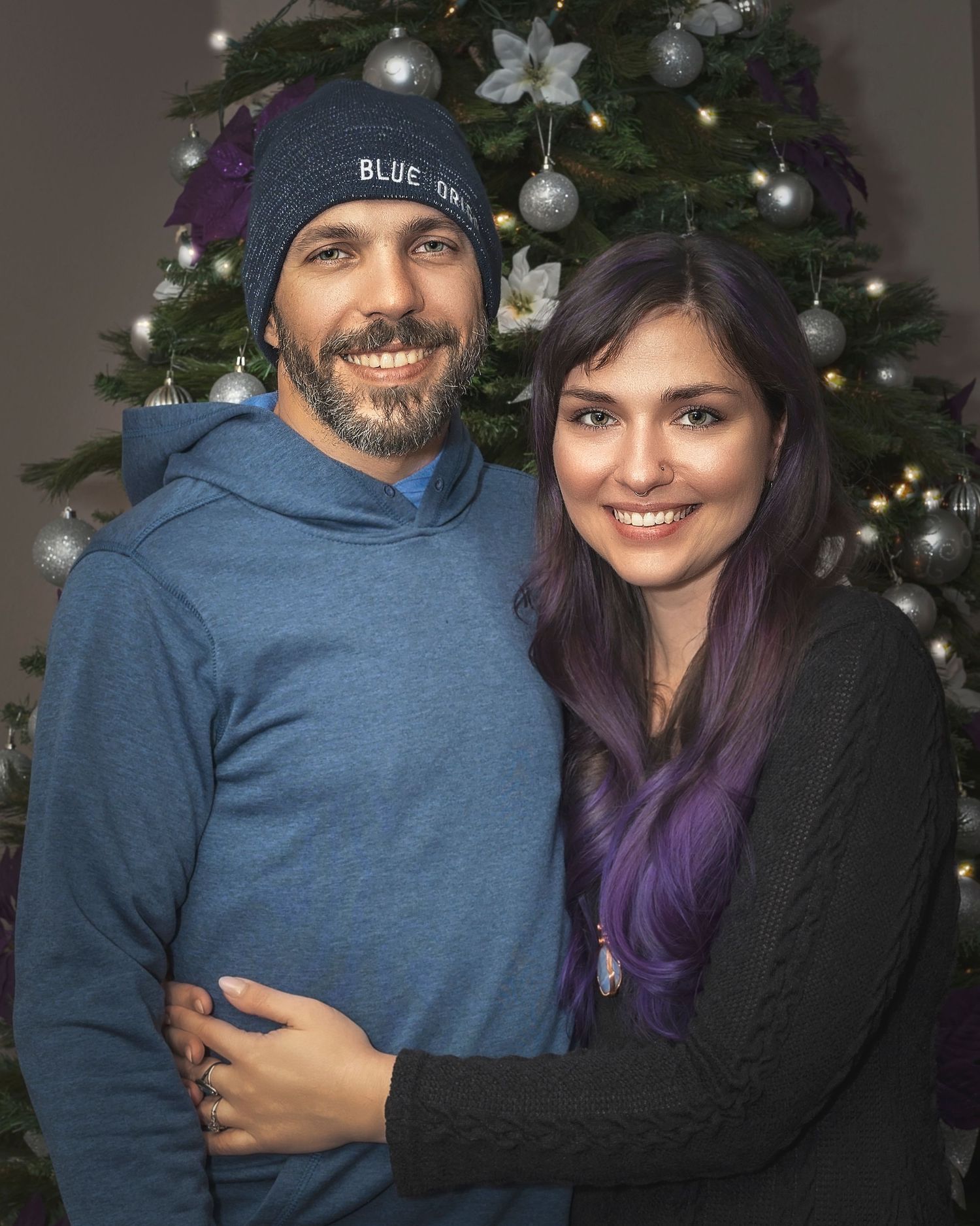 Couple smiling together in front of a decorated Christmas tree, woman with purple hair, man wearing a blue beanie hat.