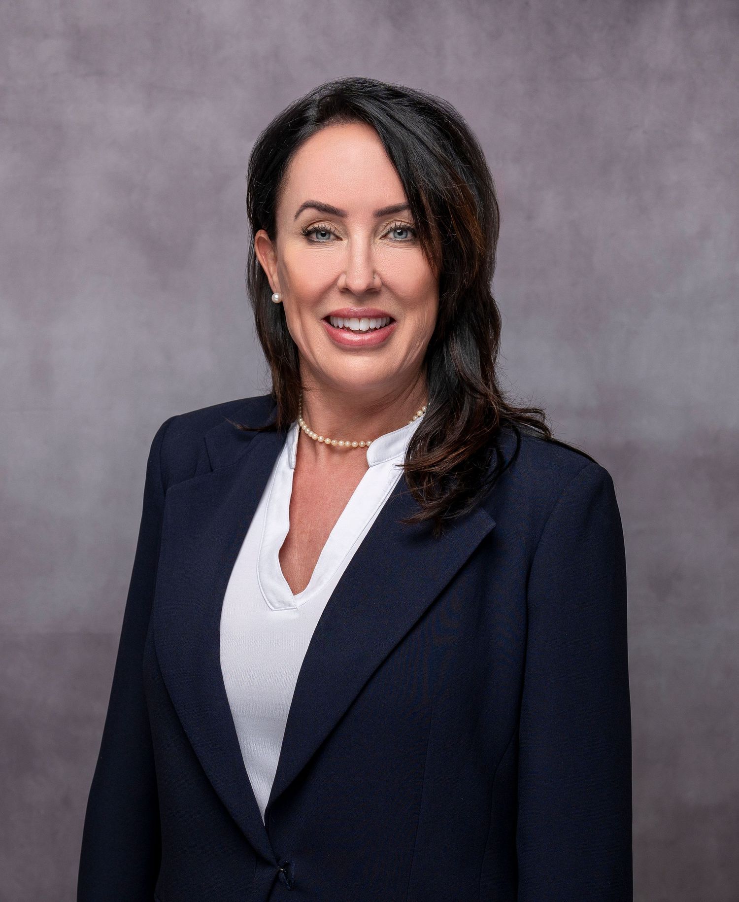 Professional woman in navy blazer and pearl necklace poses for formal business headshot against gray background.