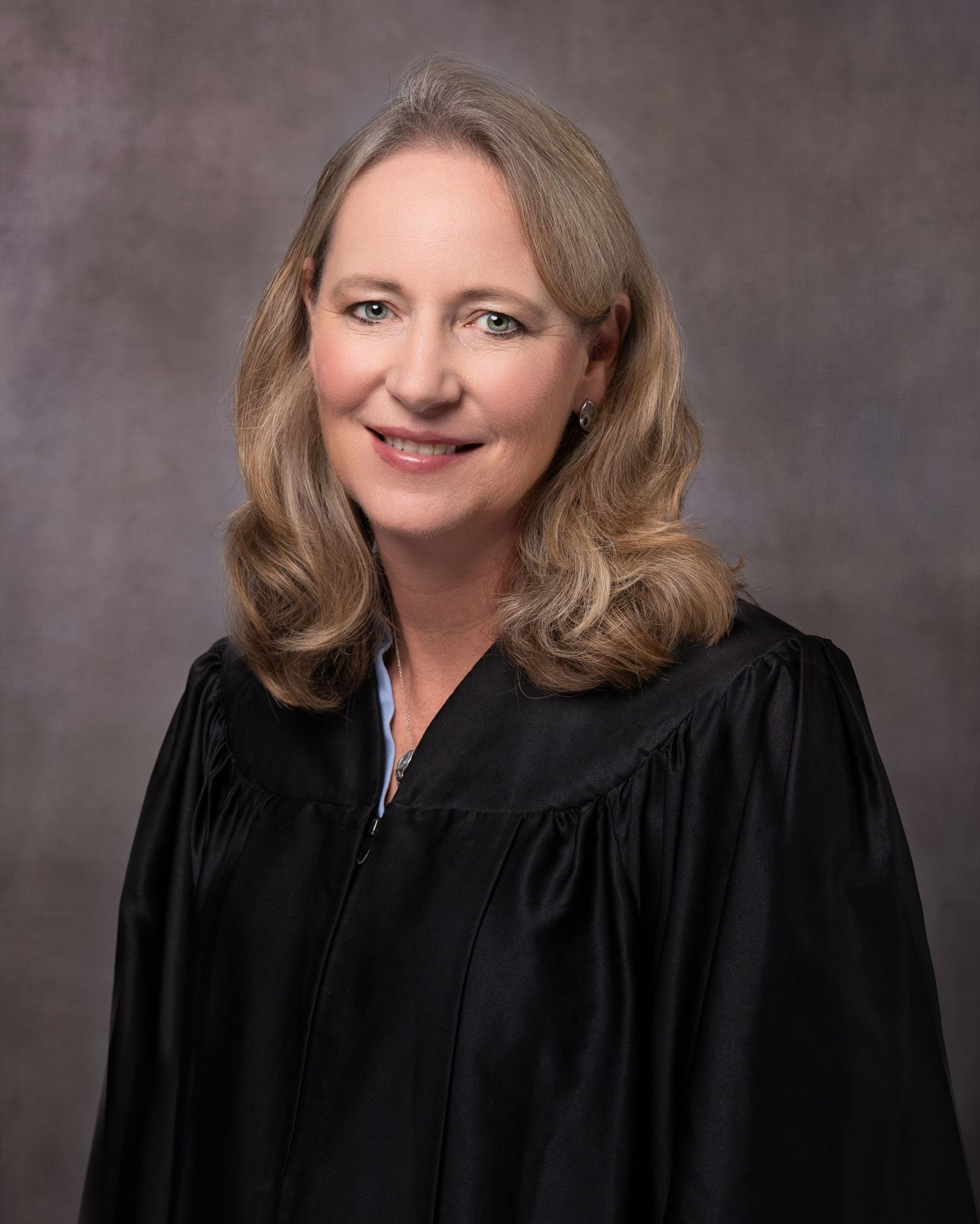 Professional portrait of a female judge wearing black judicial robes, smiling against a gray studio background.