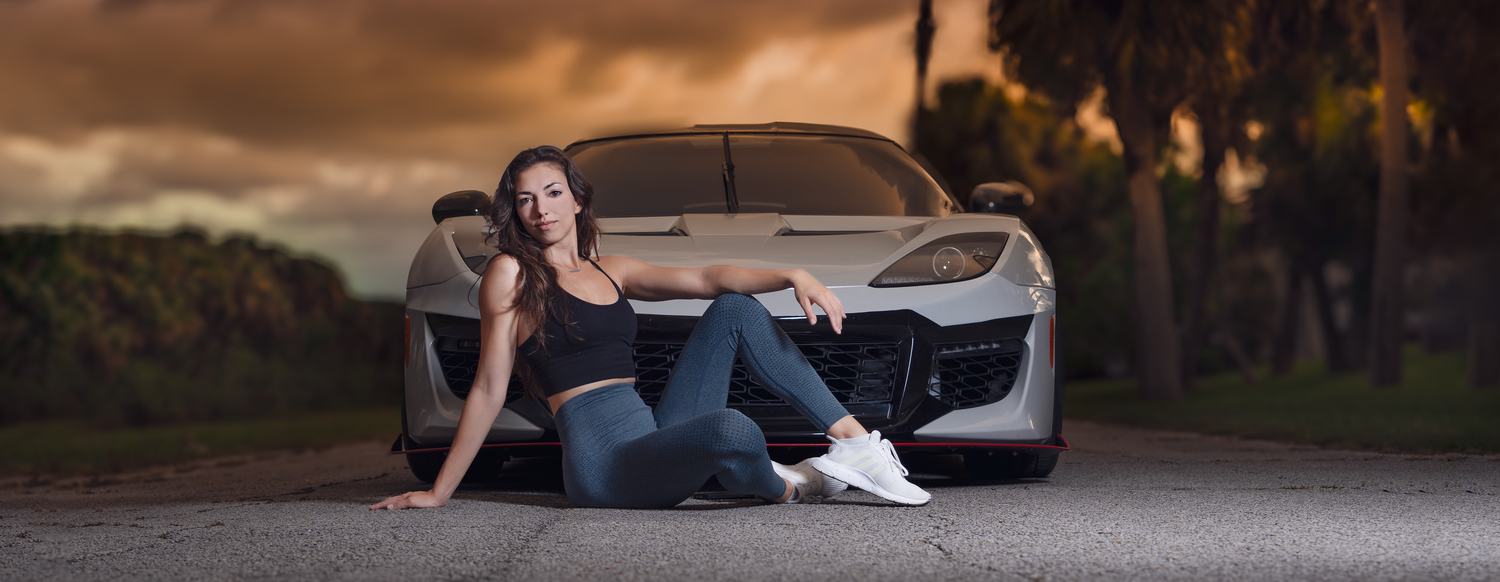 Young woman in jeans poses in front of a sleek sports car at sunset with dramatic cloudy skies.