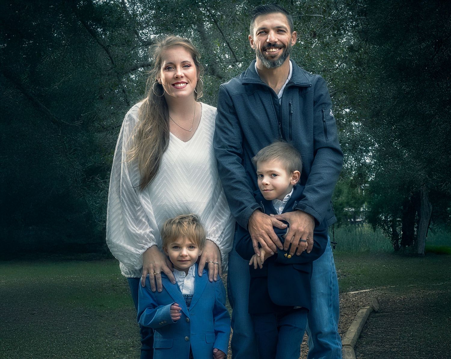 Family portrait of parents with two young boys in blue suits posing outdoors in a park setting.