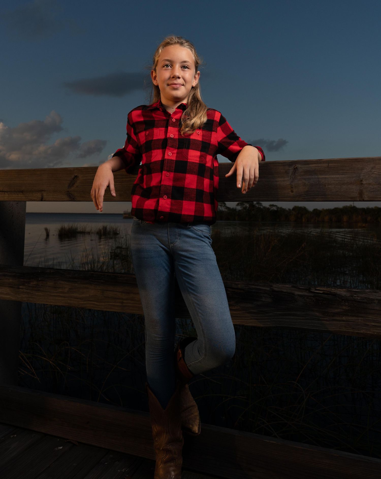 Young girl in red plaid shirt and jeans leaning on a wooden fence at sunset.
