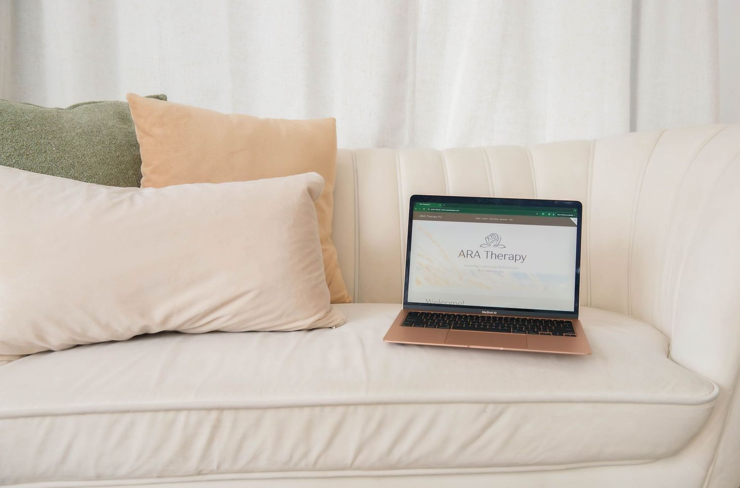 Rose gold laptop showing ARA Therapy website displayed on a white couch with pillows.