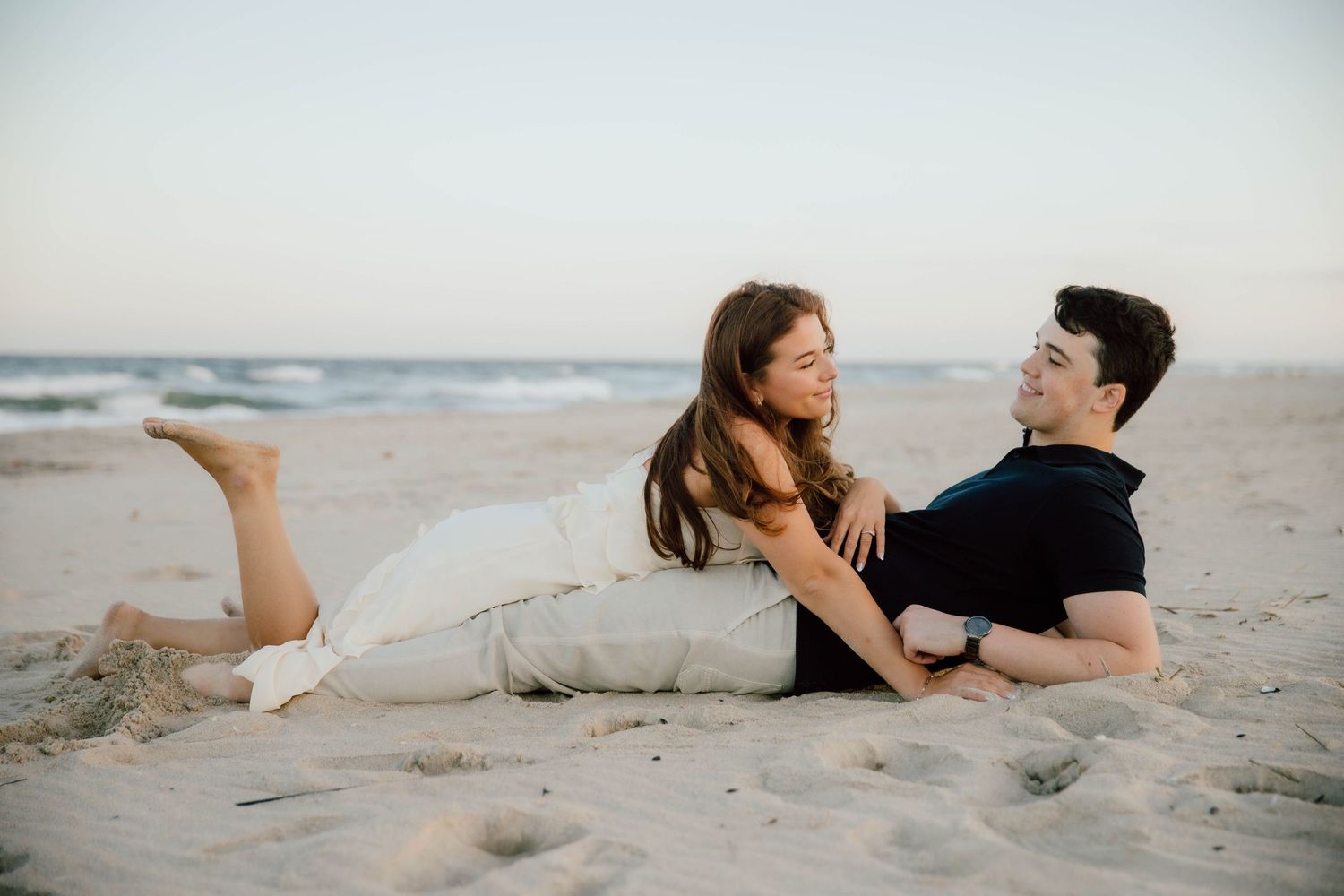 Couple relaxing together on a sandy beach at sunset, smiling and gazing at each other.