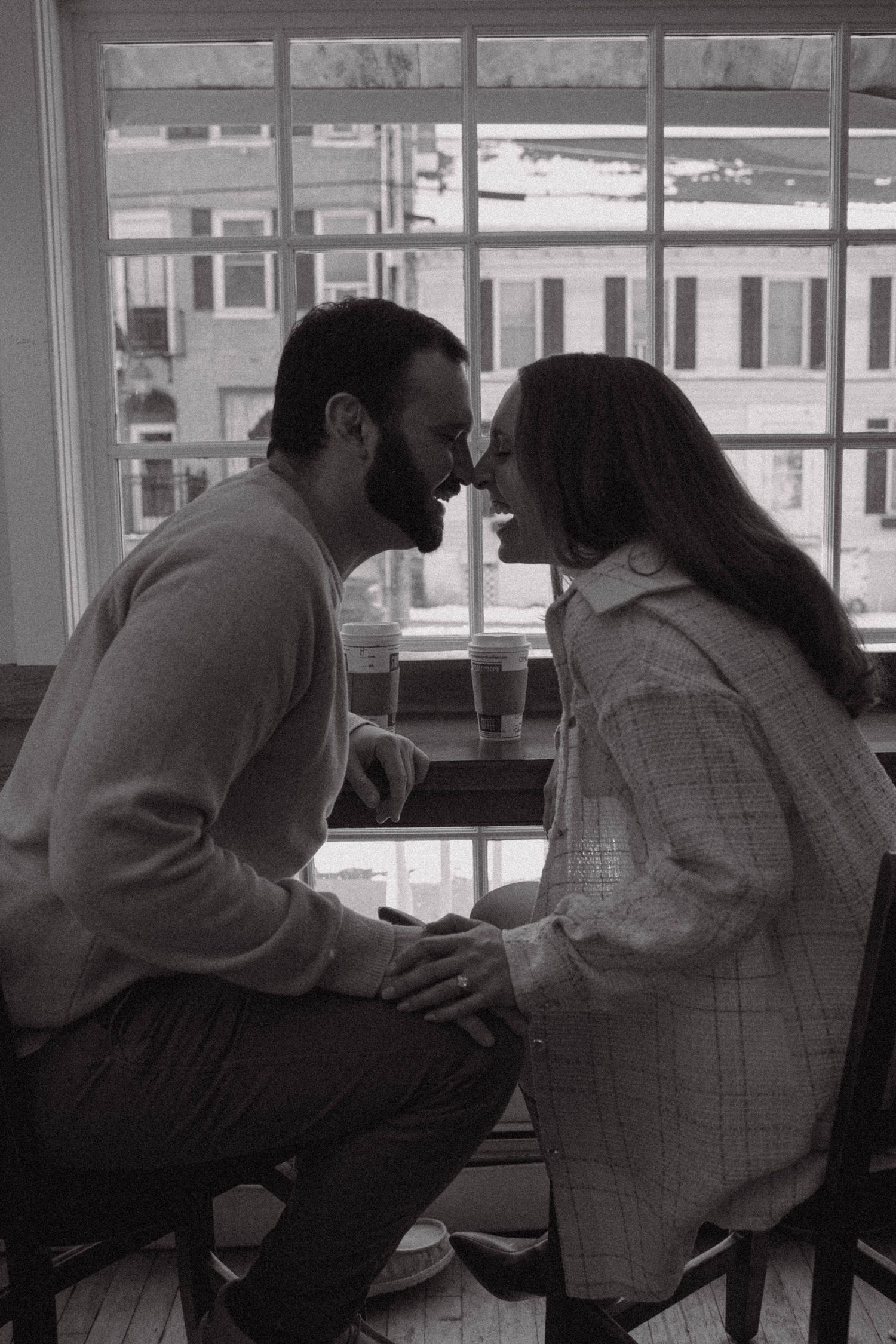 Black and white photo of a couple leaning in to kiss by a large window indoors.