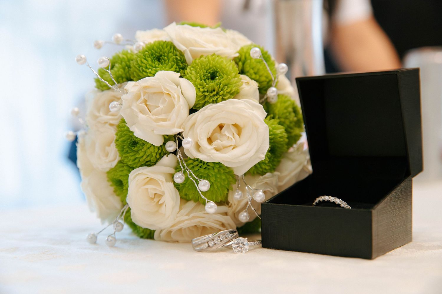 Elegant wedding bouquet of white roses and green chrysanthemums with pearl accents beside a diamond ring in a black box.