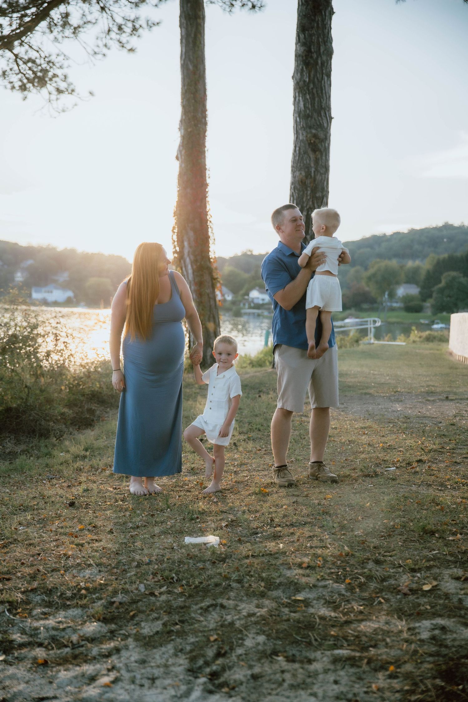A pregnant woman in a blue dress stands with her husband holding a baby and a toddler near a lakeside at golden hour.