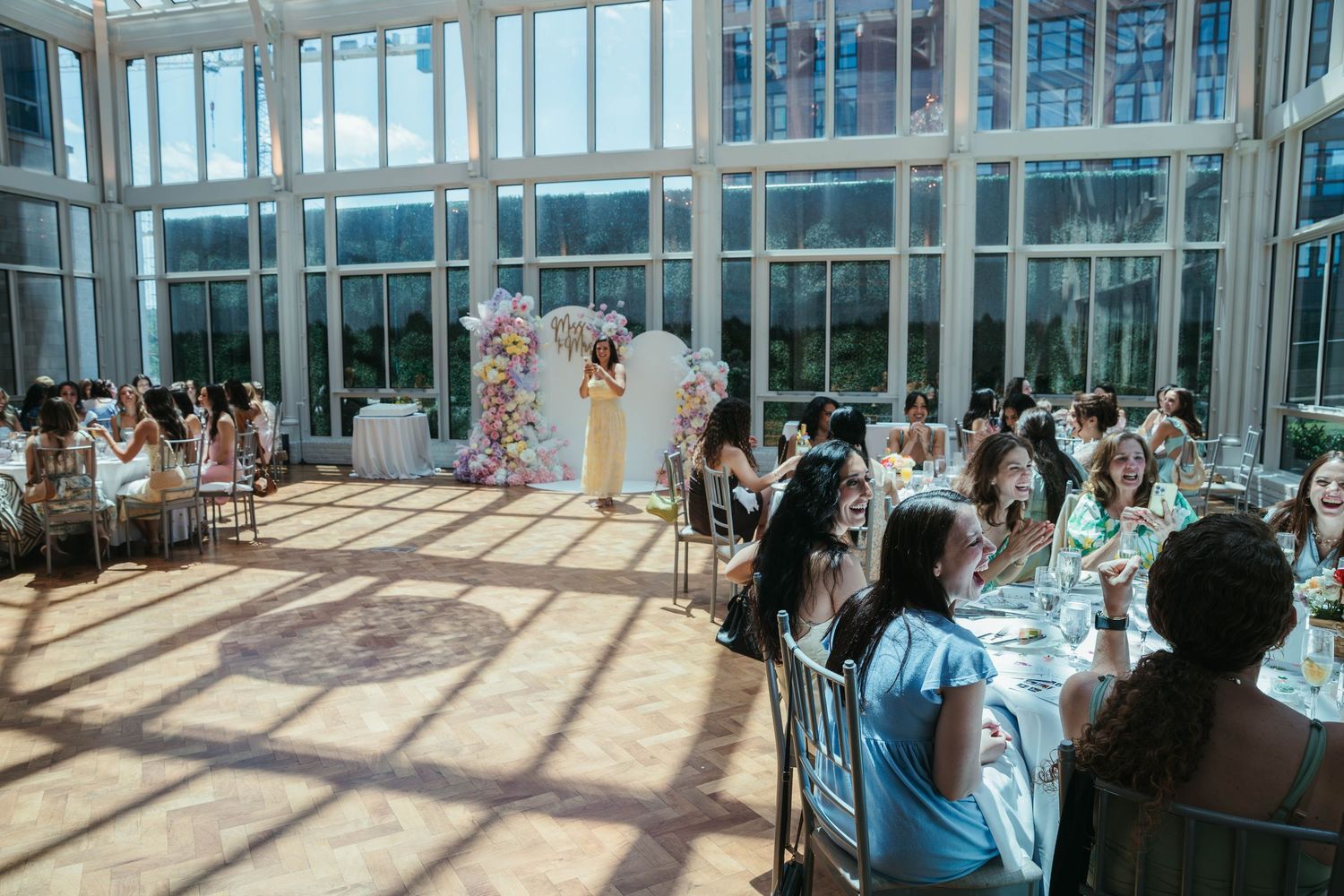 Elegant indoor wedding reception in a sunlit glass conservatory with floral arch and guests seated at decorated tables.