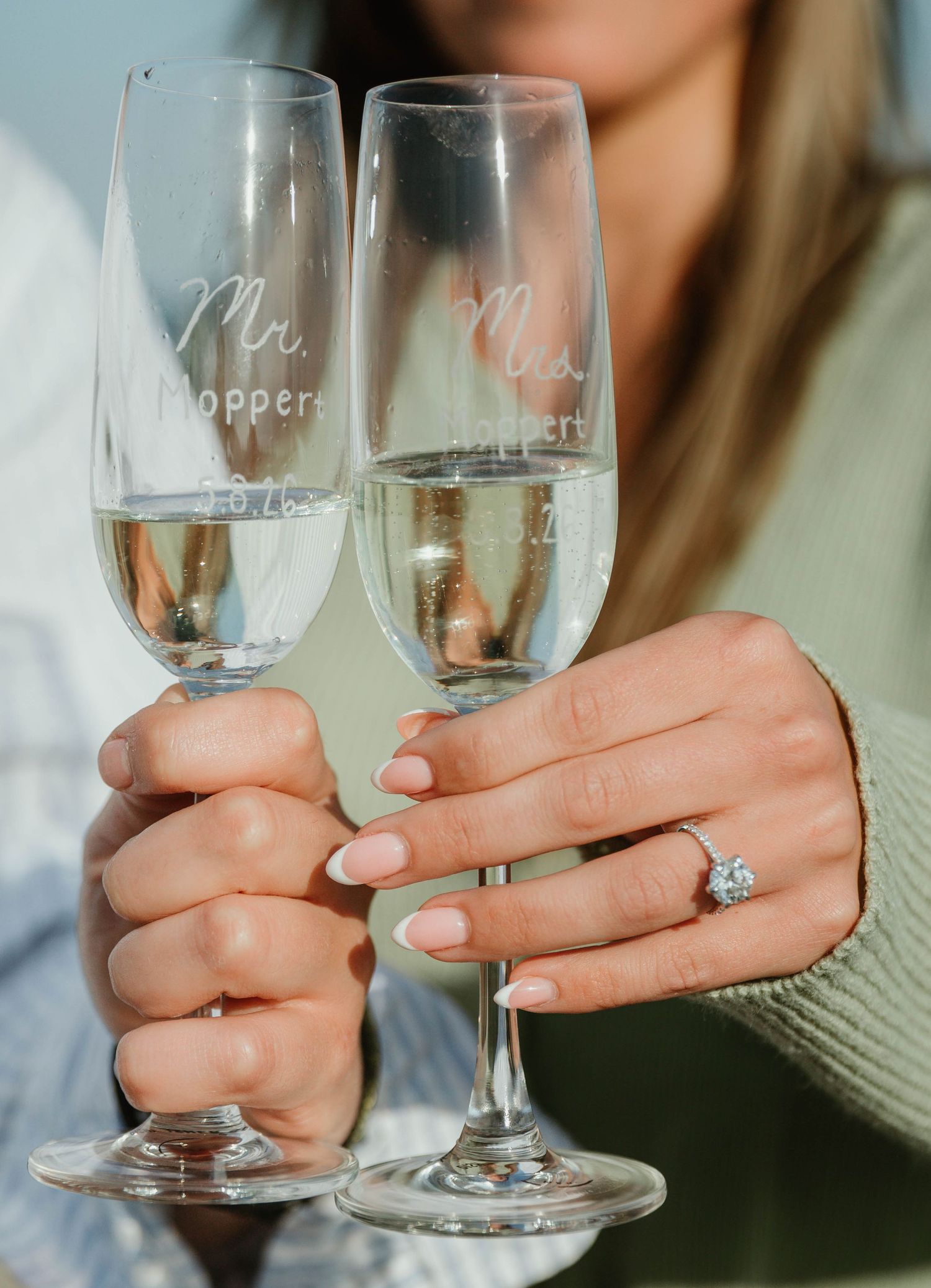 Hands holding engraved Mr. and Mrs. champagne flutes with engagement ring visible on woman's finger.