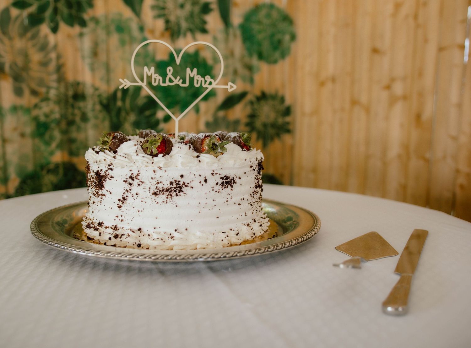 White wedding cake with Mr & Mrs topper on silver plate, decorated with chocolate shavings and berries, with cake server beside it.