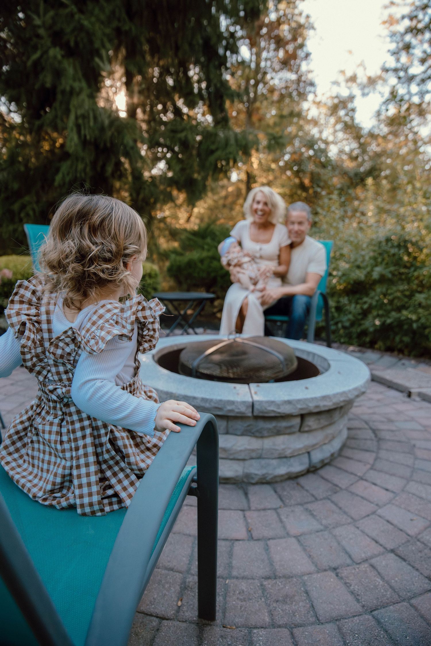 Young child in plaid dress sits by stone fire pit while grandparents relax together in a cozy backyard garden setting.