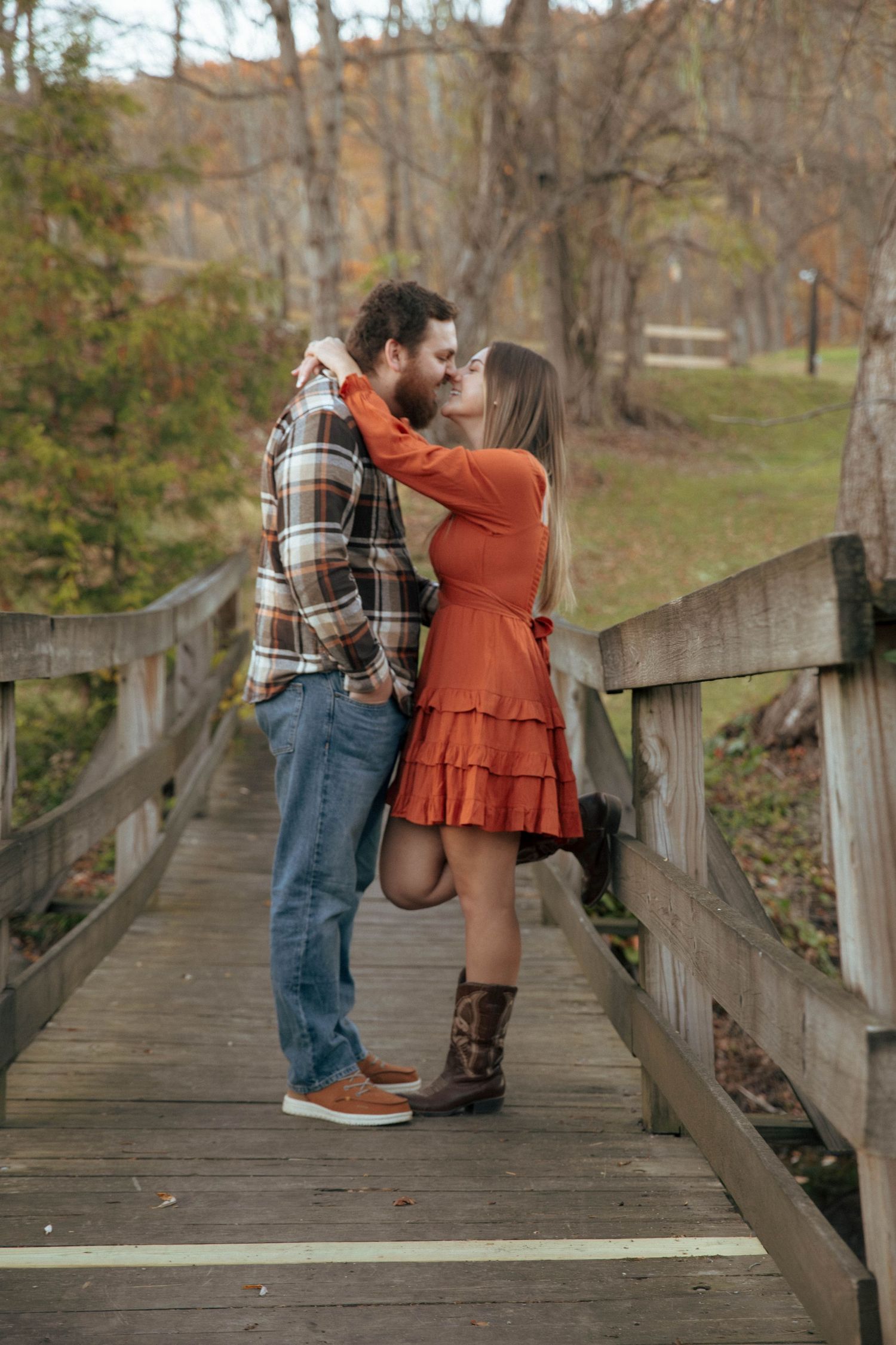 Couple sharing romantic kiss on wooden bridge surrounded by colorful autumn foliage in park.