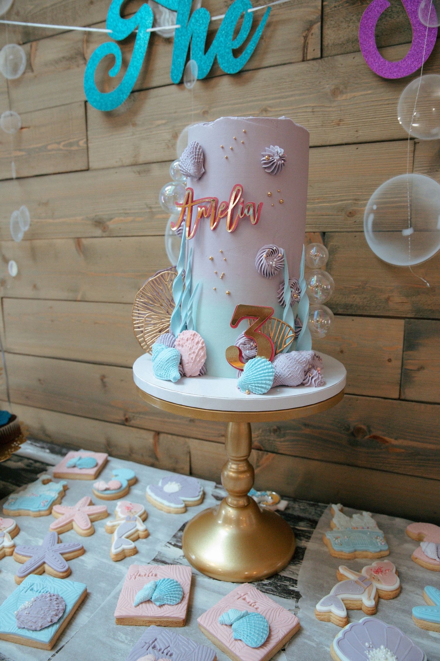Elegant mermaid-themed birthday cake with seashells and name Amelia on gold cake stand with cookies.