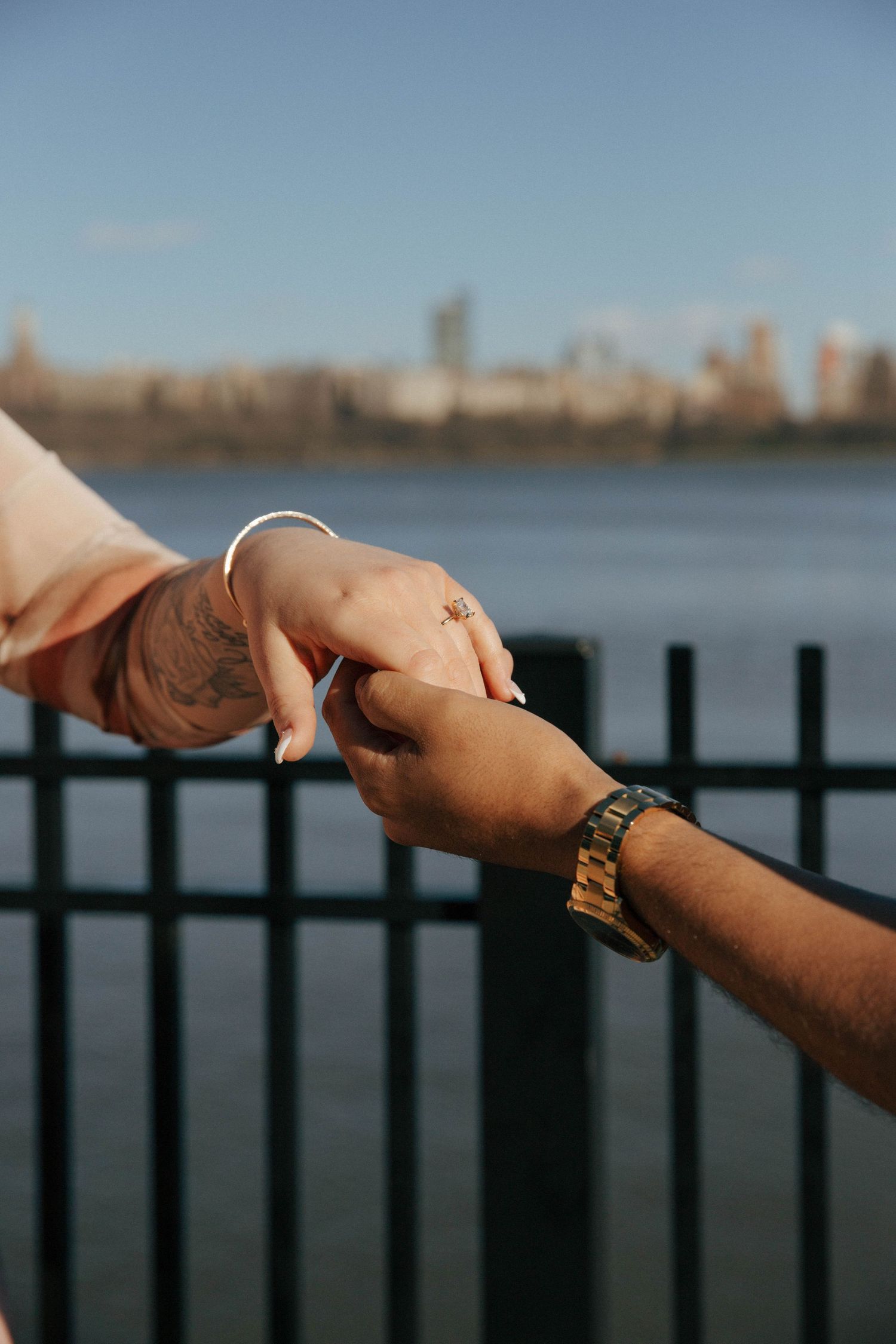 Close-up of an interracial couple holding hands near a waterfront fence, woman wearing an engagement ring.