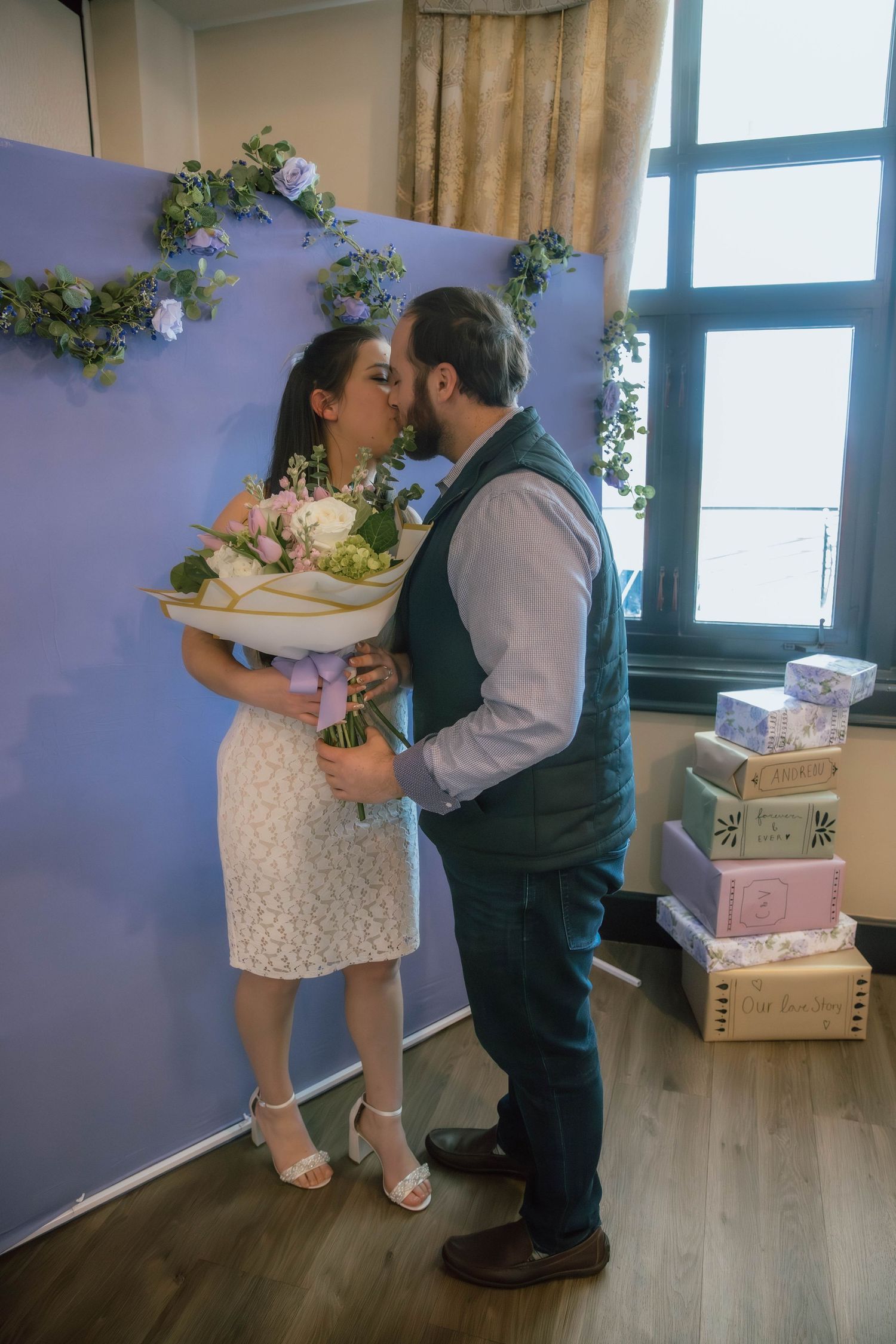 A couple shares a romantic kiss, the woman in a white dress holding a bouquet of flowers in a decorated indoor setting.