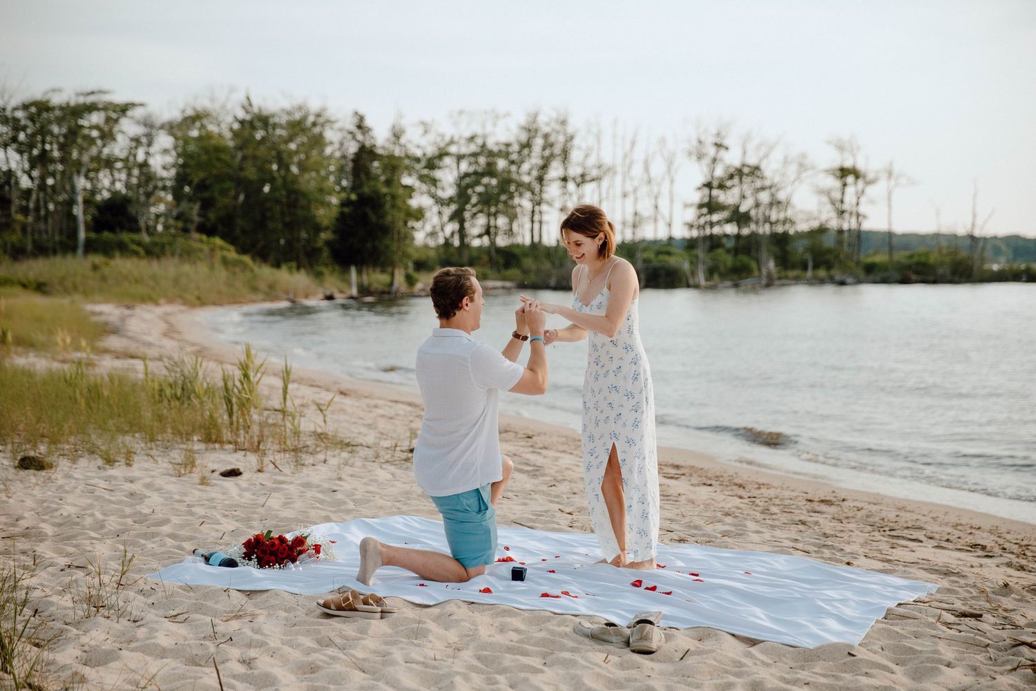 Beach proposal at sunset with man on one knee presenting ring to woman in white dress.