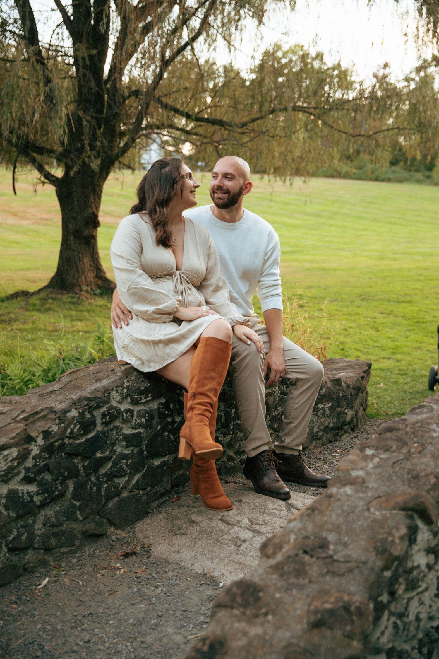 A couple sits on a stone wall in a park, smiling at each other, woman in cream dress and brown boots.