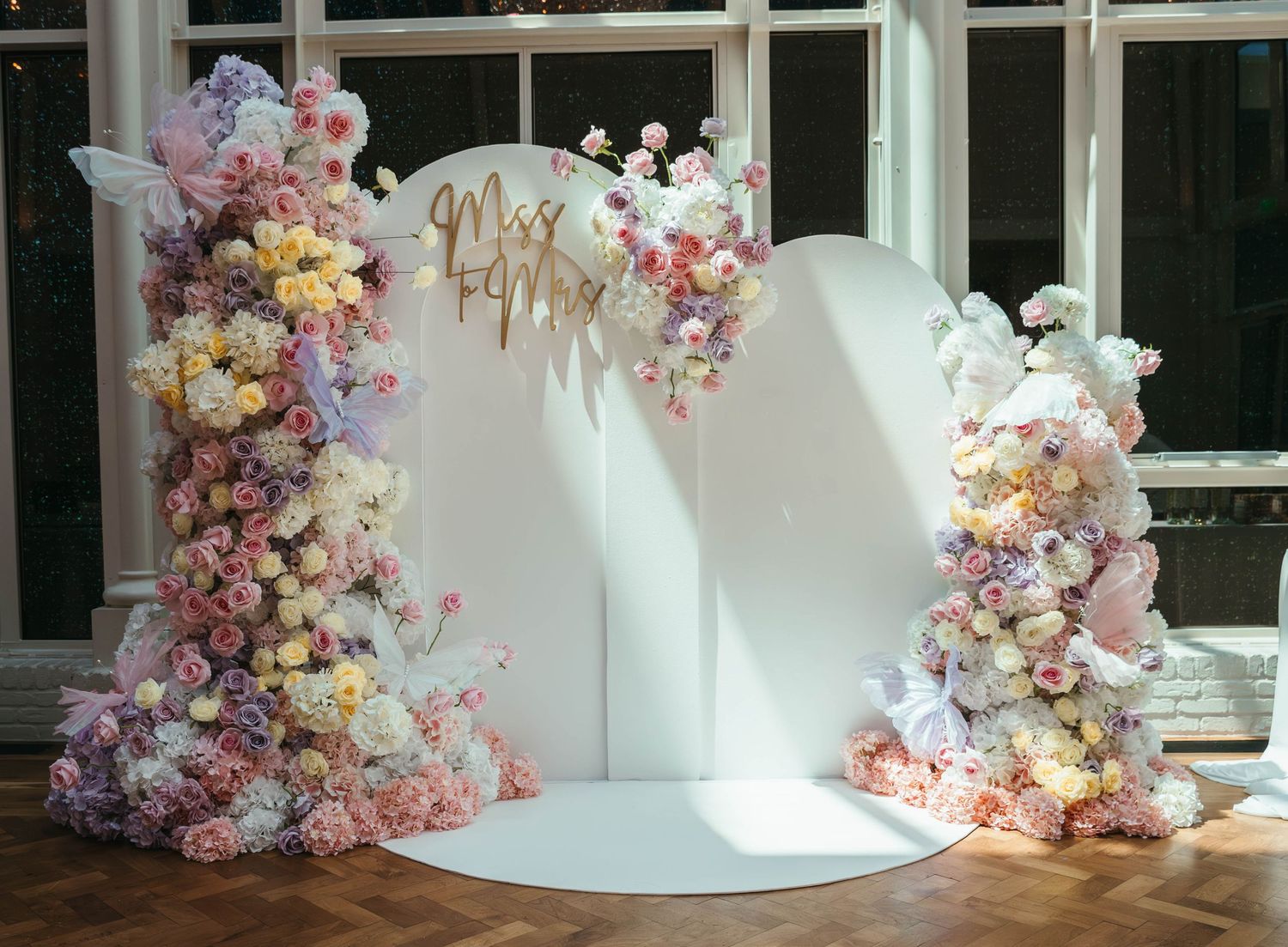 Elegant wedding arch adorned with pink, white, and purple floral arrangements on a circular white backdrop with gold Mr & Mrs sign.