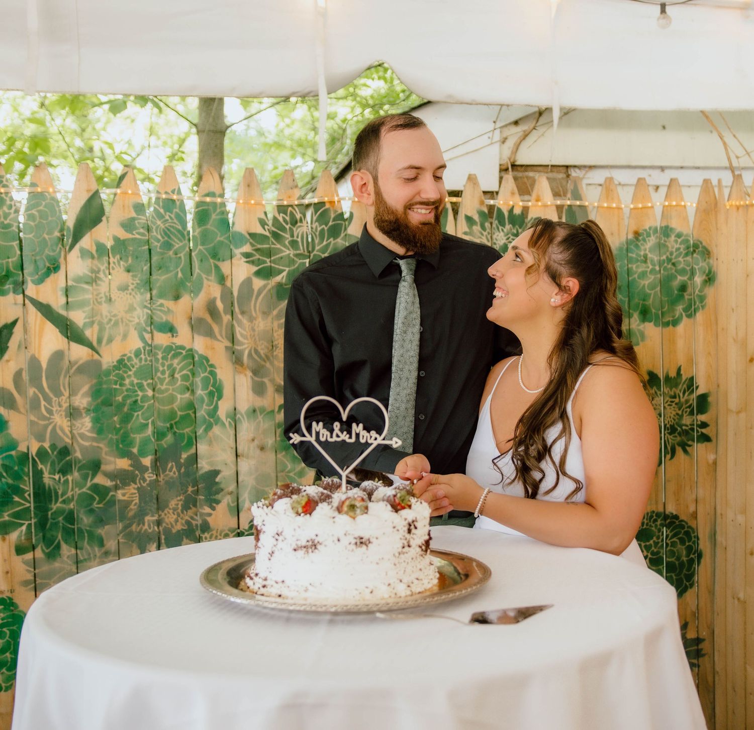 Newlywed couple smiling at each other while cutting their white wedding cake at an outdoor garden reception.