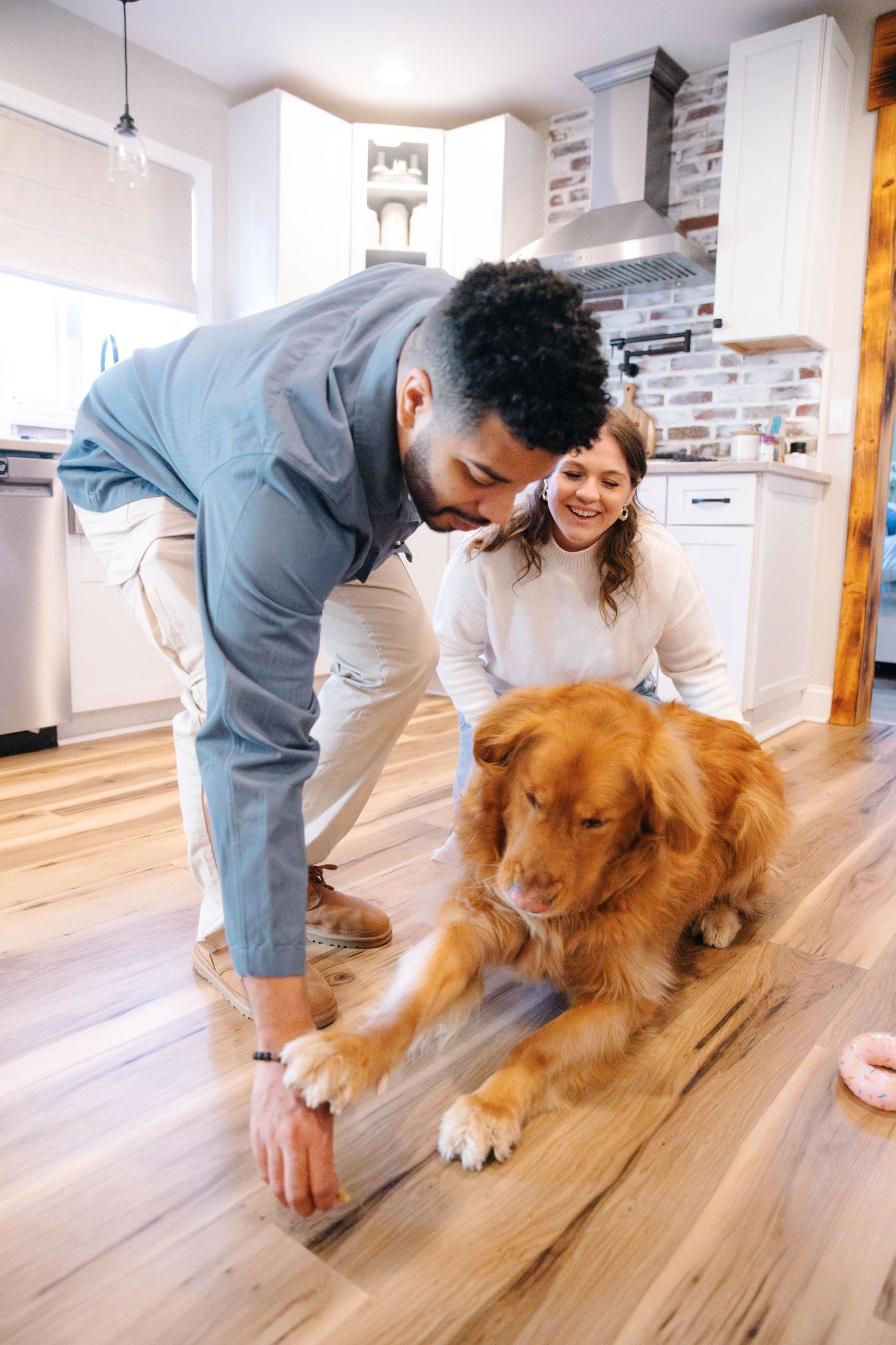 Couple playing with golden retriever dog on hardwood floor in modern kitchen.