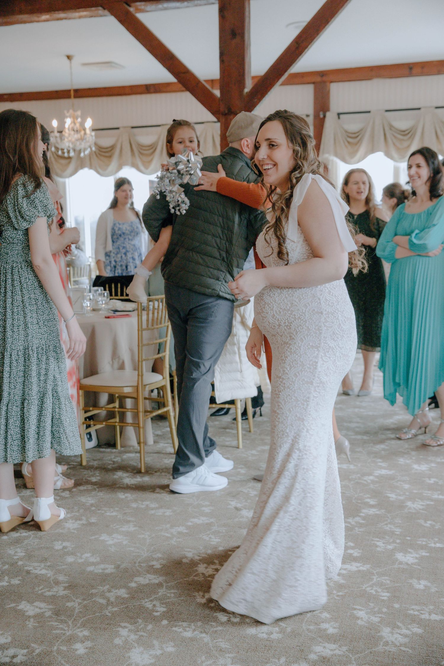 A smiling bride in a white lace gown dances with a guest at her wedding reception surrounded by attendees.