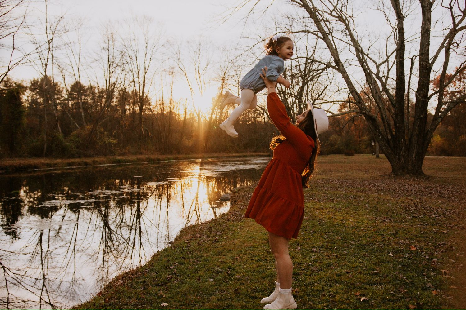 A mother lifts her toddler at golden hour by a serene pond, surrounded by autumn trees in a park.
