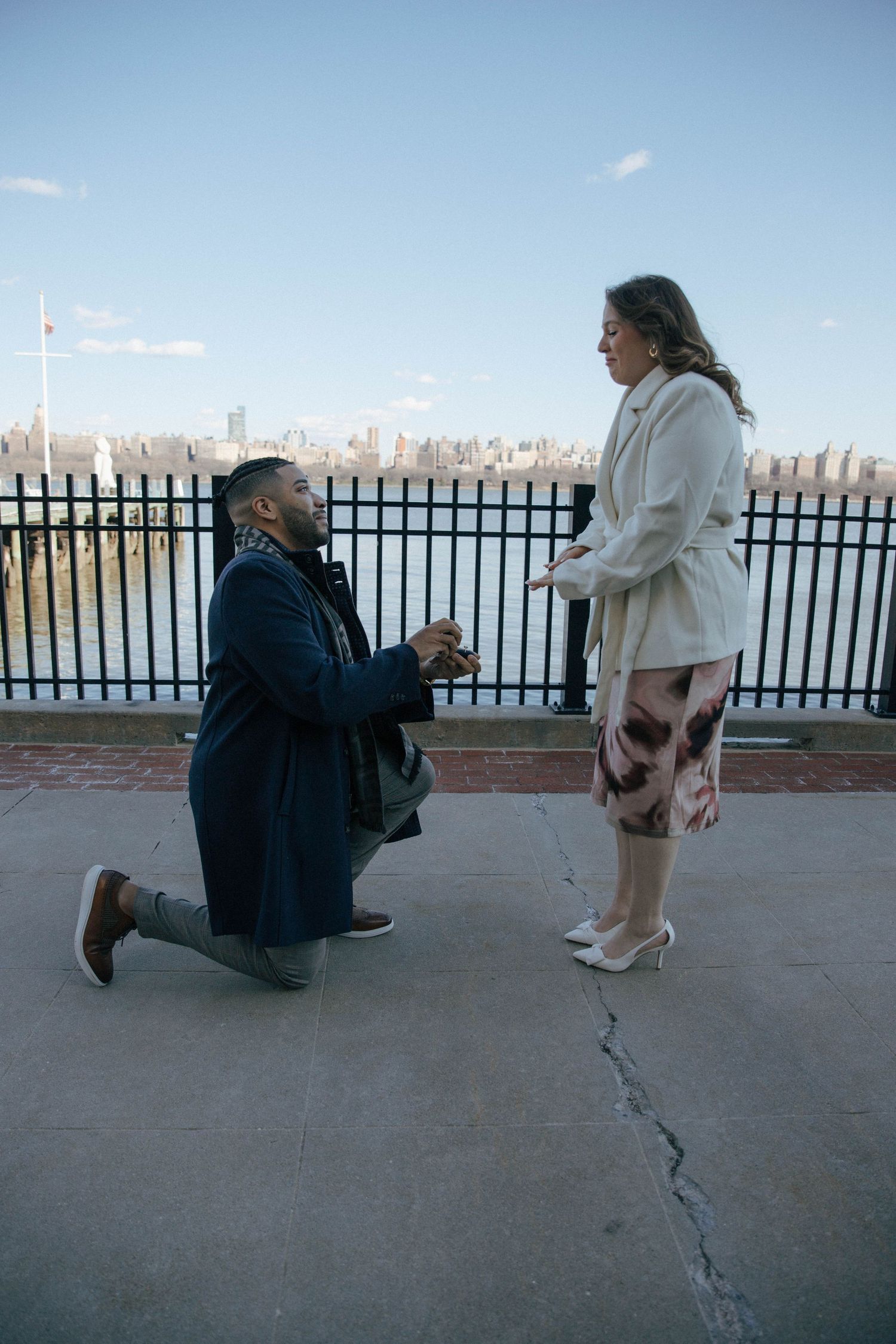 Man on one knee proposing to a woman on a waterfront promenade with a city skyline in the background.