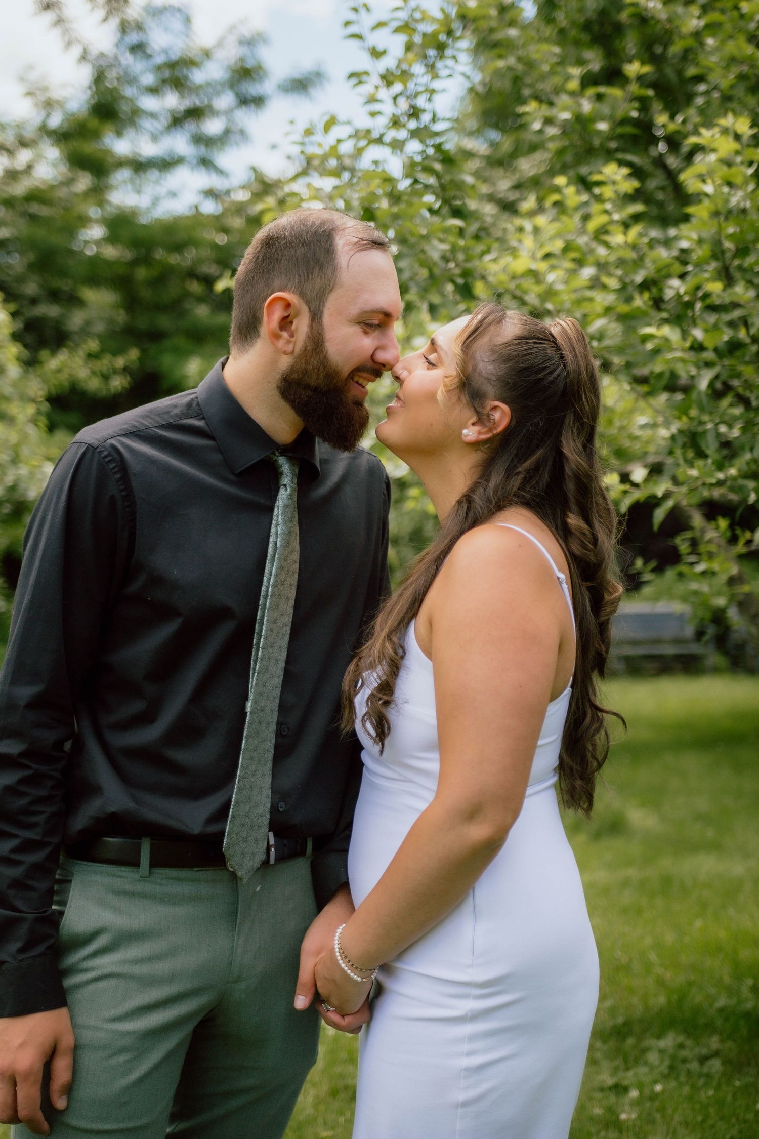 Bearded man in black shirt and woman in white dress share tender moment in green garden setting.