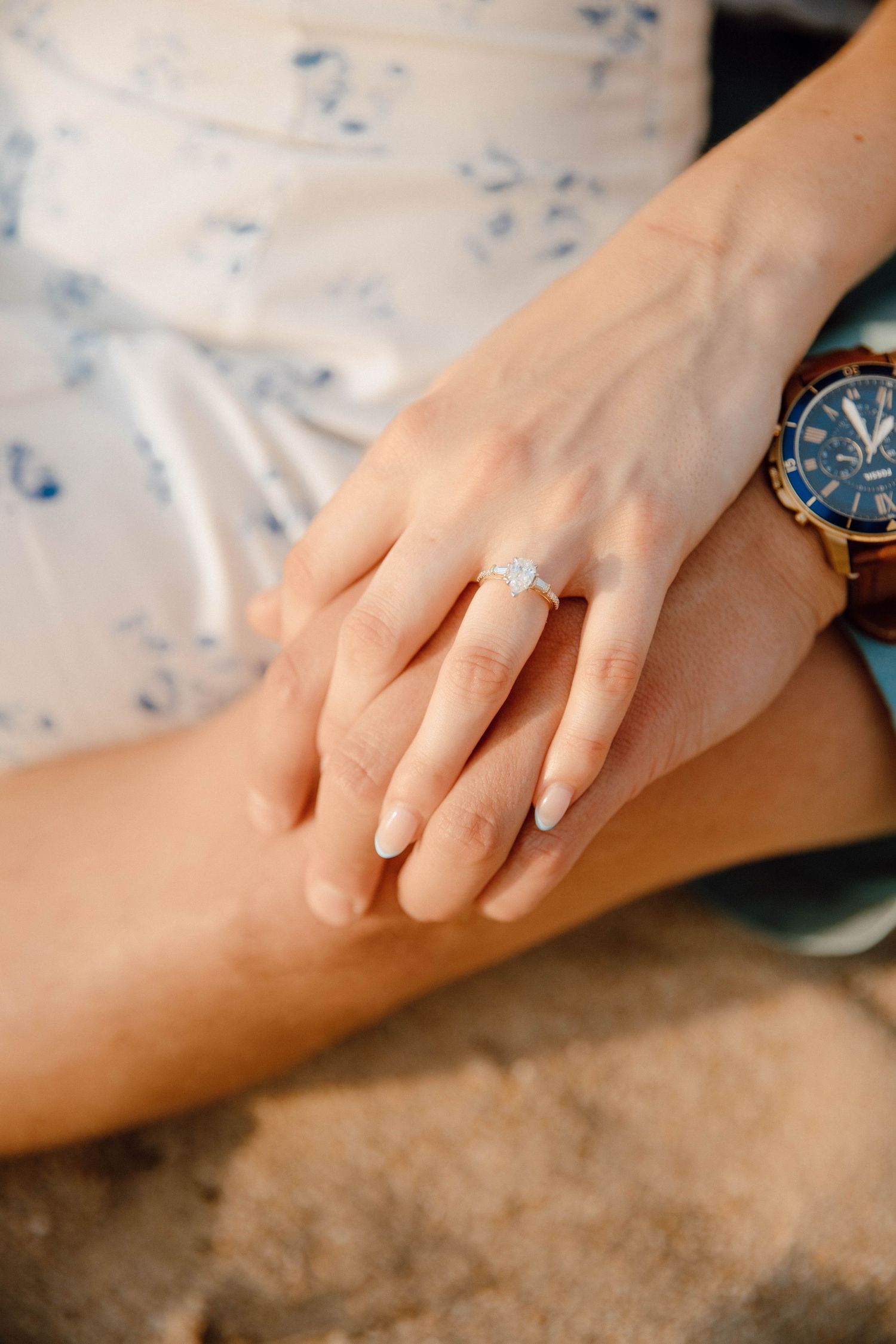 Close-up of a woman's hand wearing a diamond engagement ring resting on her lap beside a watch.