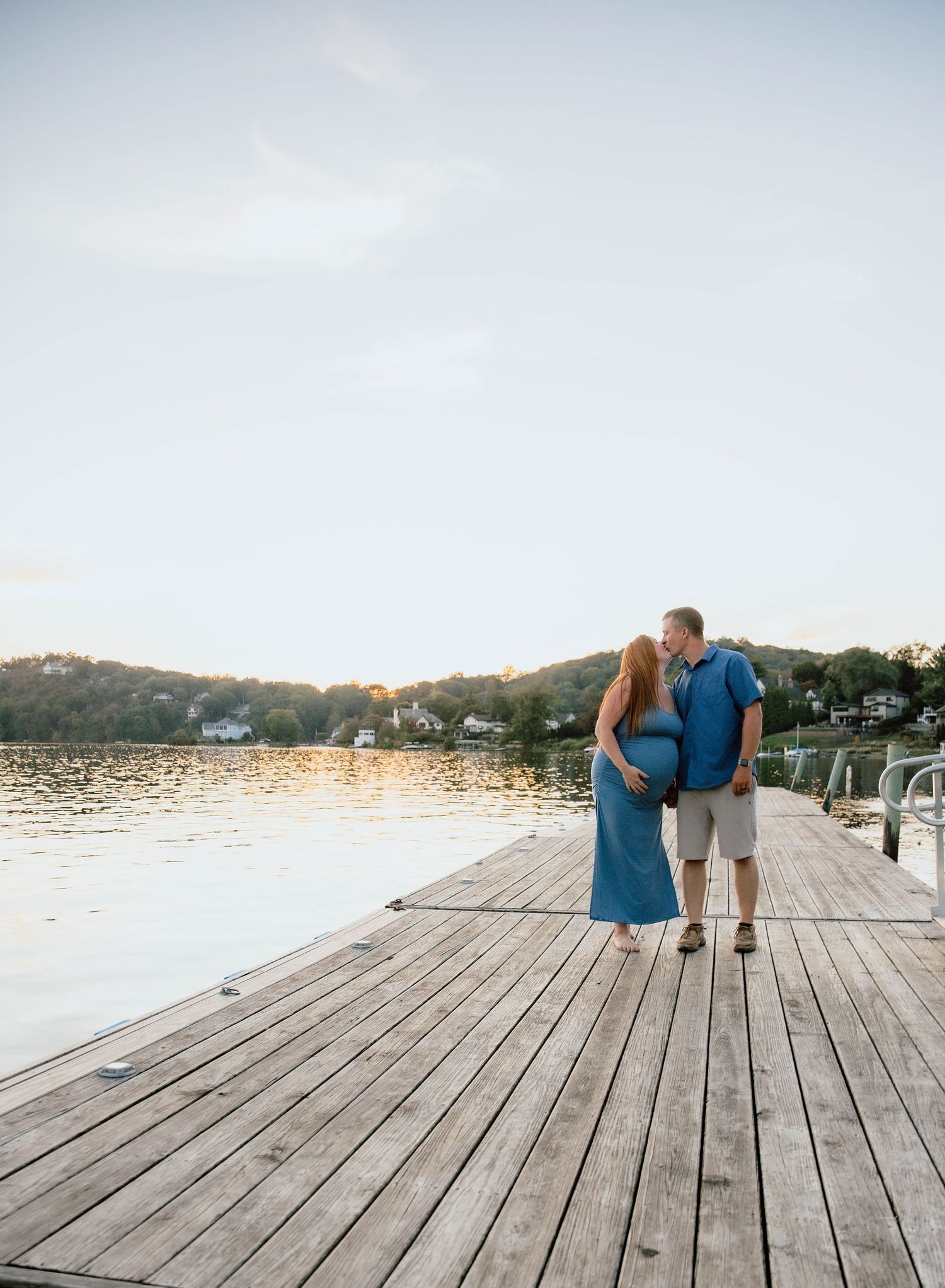 Pregnant couple standing on a wooden dock at sunset, embracing by a calm lake with lush green hills in the background.