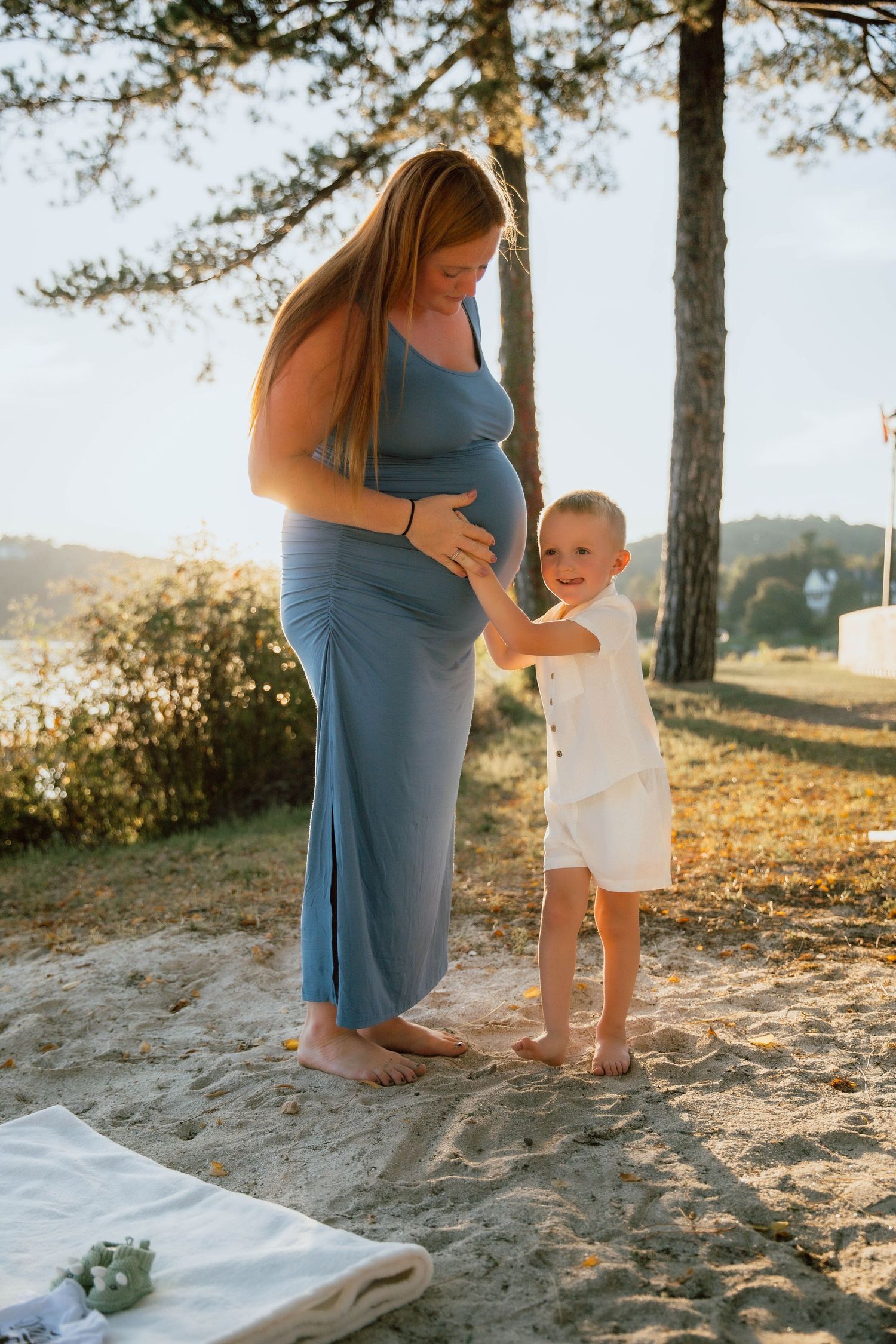 Pregnant woman in blue dress with toddler boy touching her baby bump outdoors in golden sunlight.