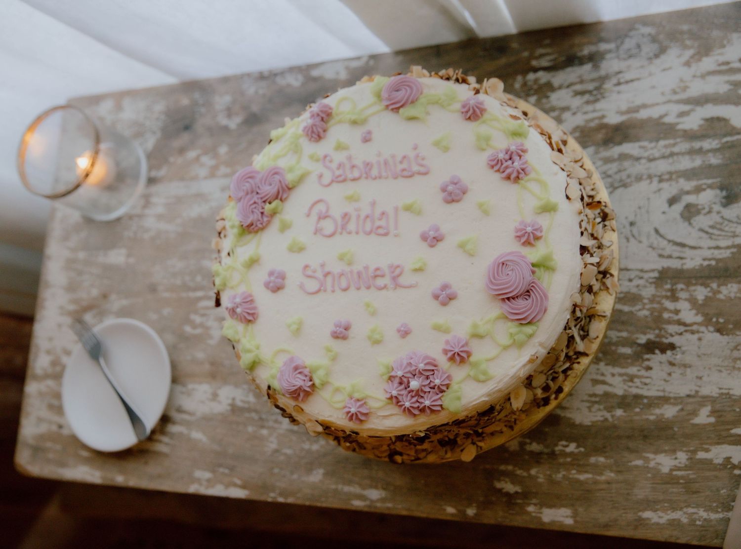 White-frosted bridal shower cake with pink floral decorations reading Sabrina's Bridal Shower on a rustic wooden table.