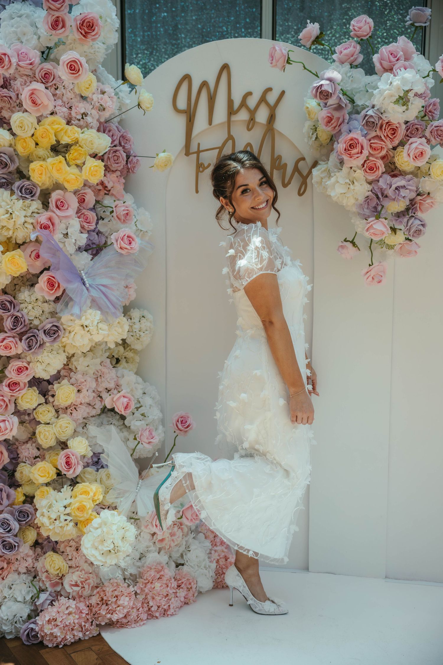 Bride in white feathered dress posing by floral arch with pastel roses and Miss sign at wedding reception.