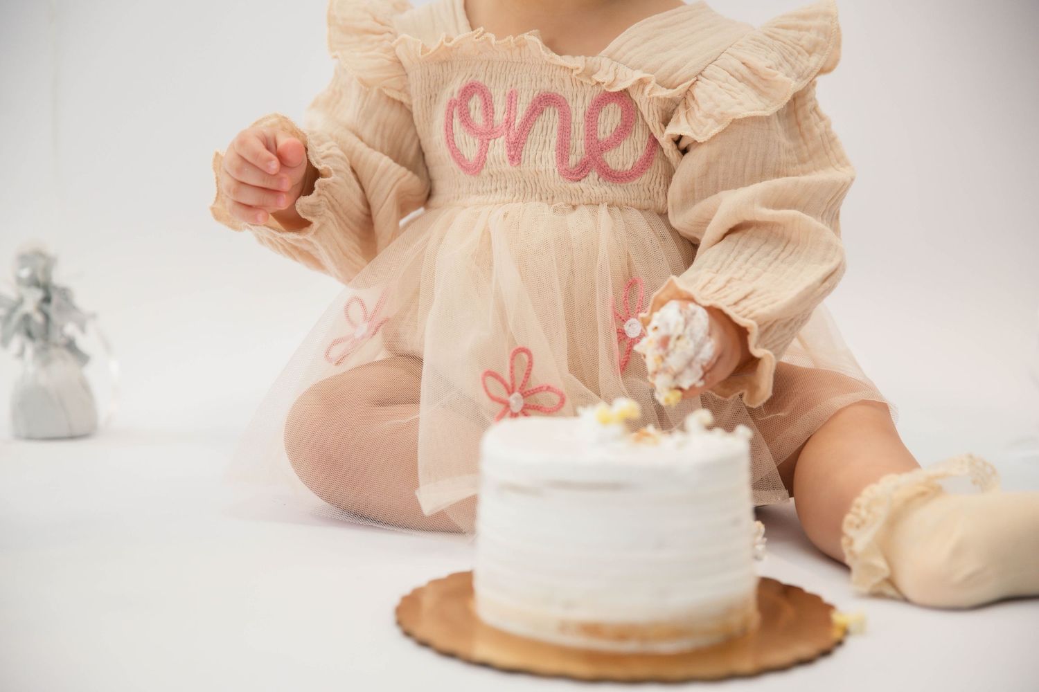 Baby girl in a beige 'one' dress smashing her first birthday white cake during a cake smash photoshoot.