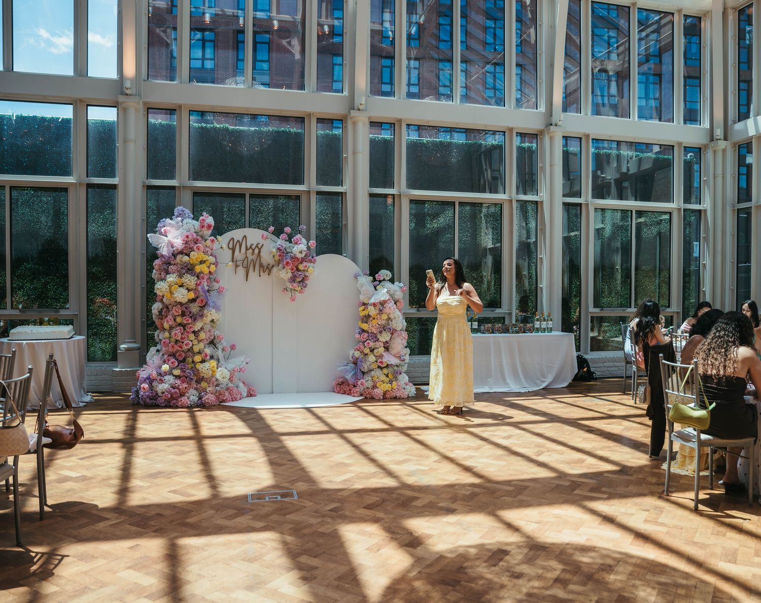 Elegant wedding venue with floor-to-ceiling windows, floral arch backdrop, and sunlit parquet flooring.