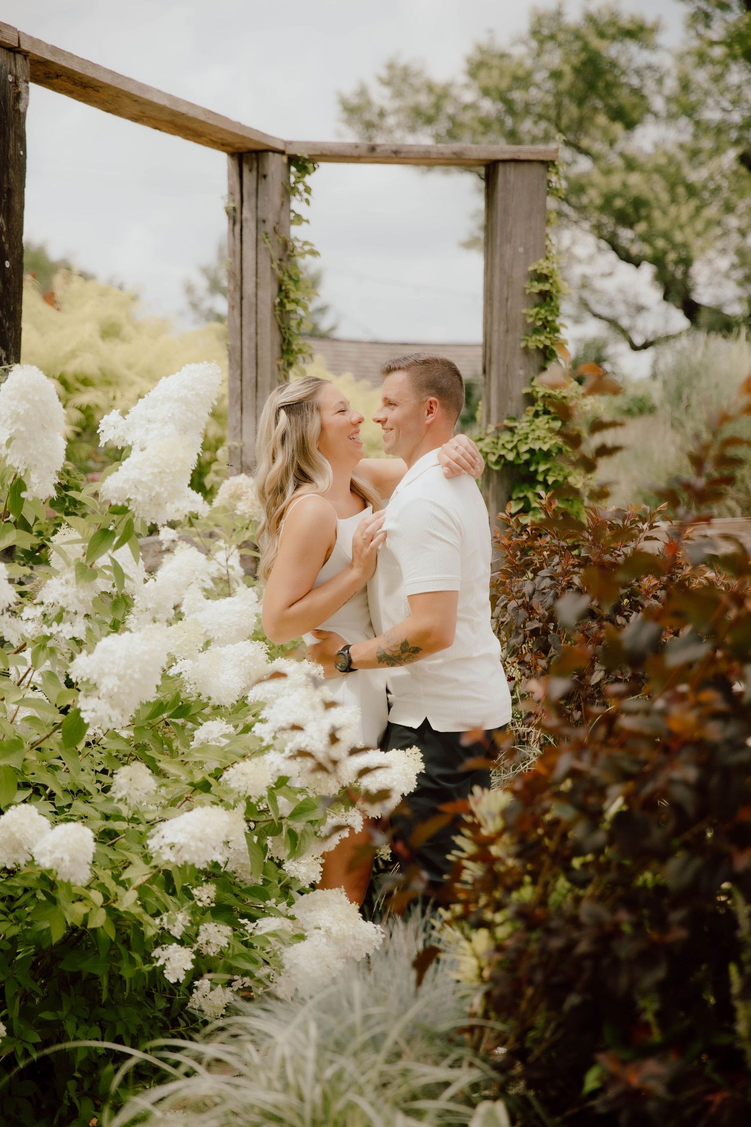 Couple embracing among white hydrangeas under a rustic wooden arbor in a lush garden setting.