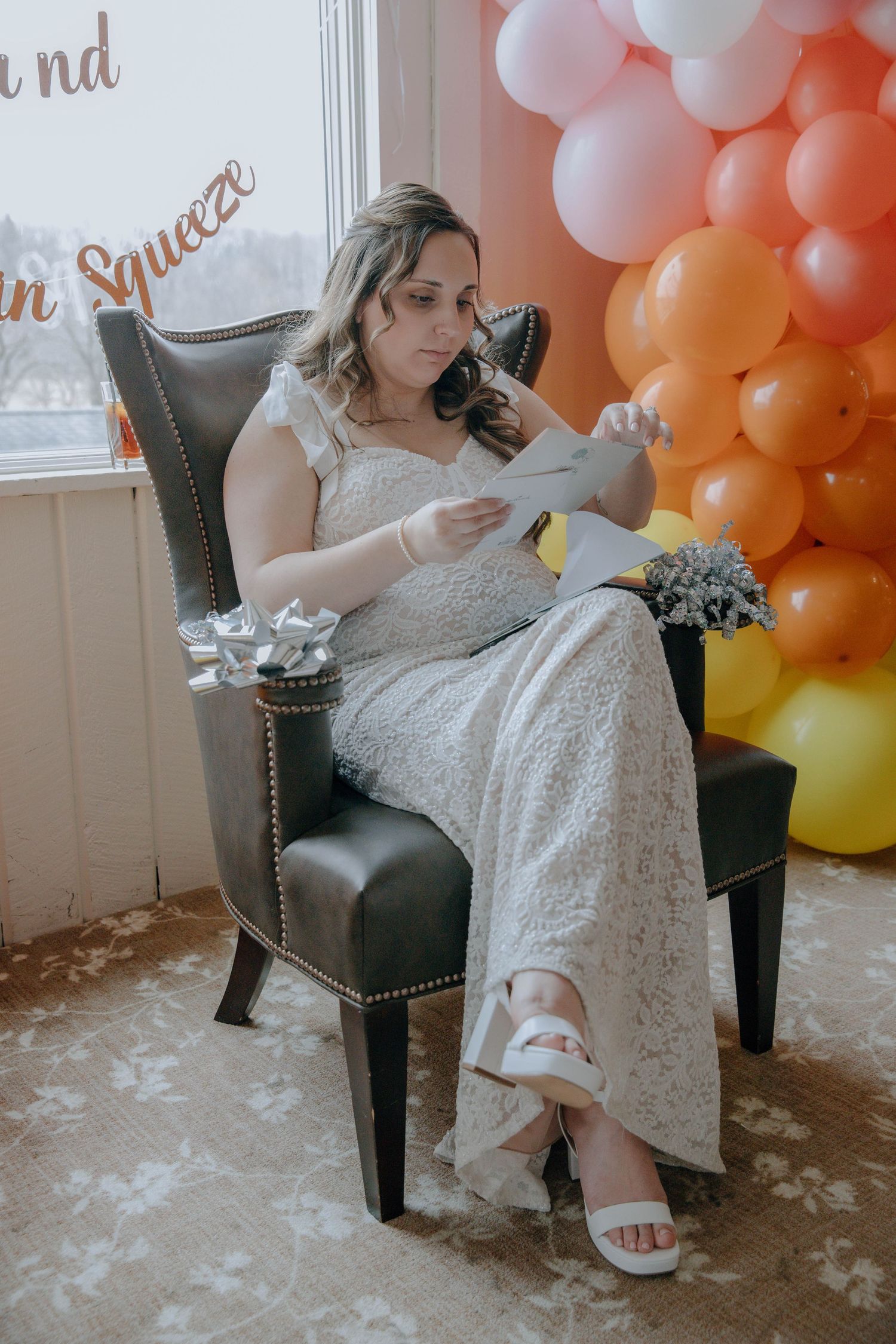 Bride in white lace gown reads a letter while seated in a leather chair, surrounded by colorful balloons.