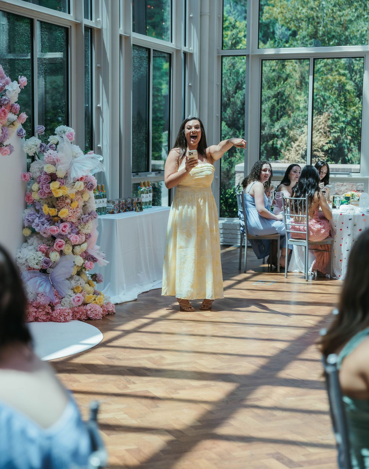 Woman in yellow dress speaks at elegant floral-decorated event venue with guests seated at tables in background.