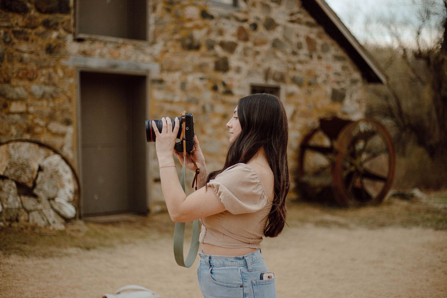 Young woman with dark hair using DSLR camera near rustic stone building with vintage wagon wheel.