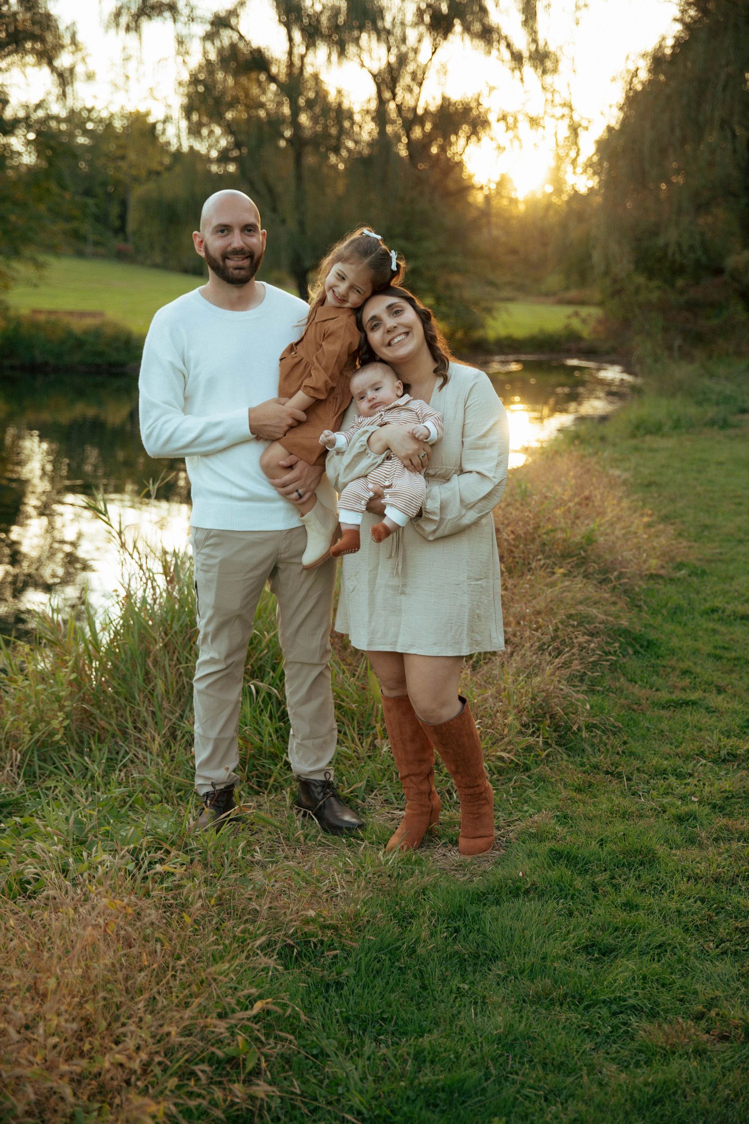 Smiling family of four posing by a pond at sunset with two young children.