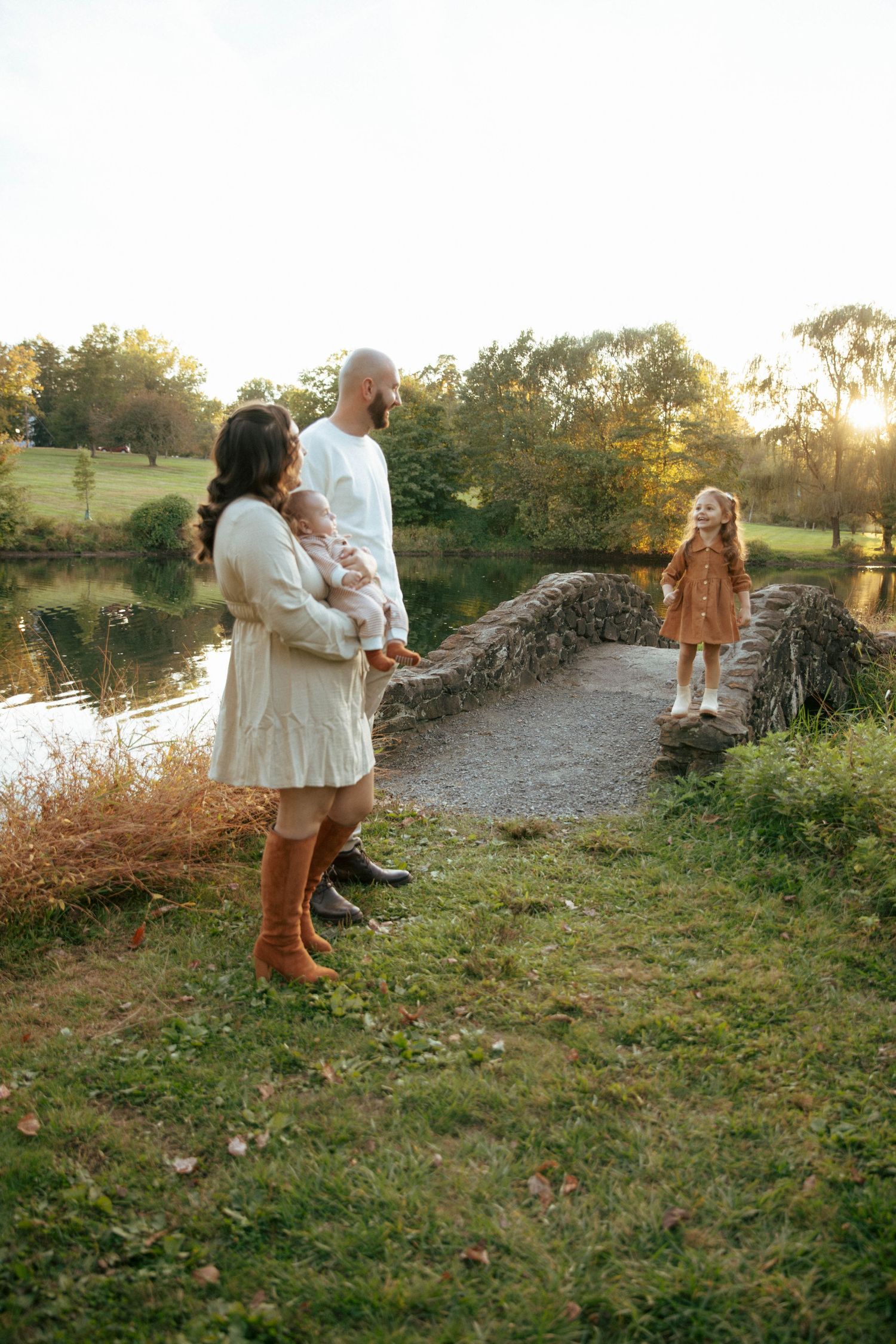 Family of four enjoying a golden sunset by a pond, parents holding baby while young girl stands on a stone bridge.